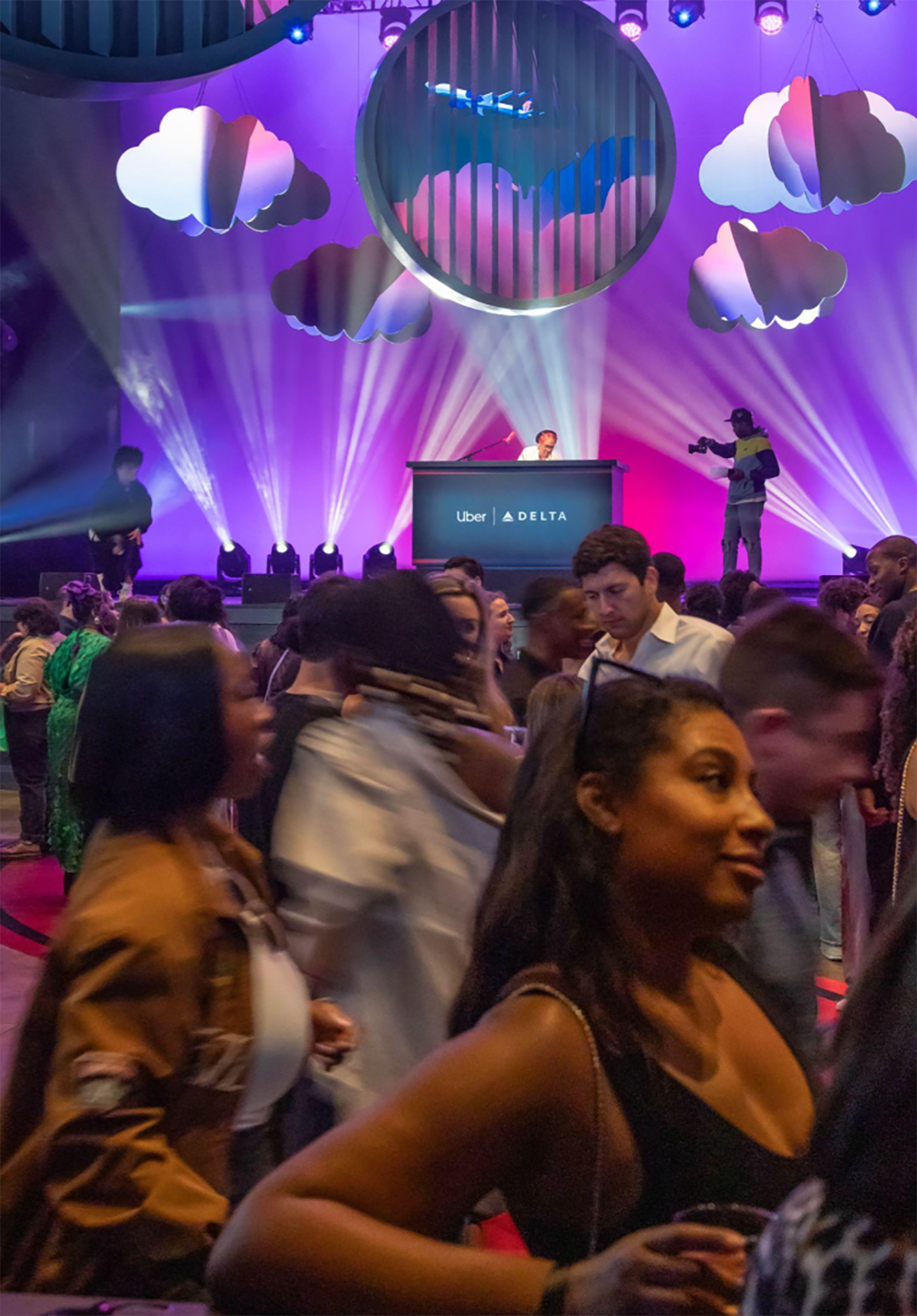 An indoor event with a crowd, a DJ booth labeled with Uber and Delta, purple and pink lighting, clouds and a circular structure with vertical bars on the stage, and people socializing.