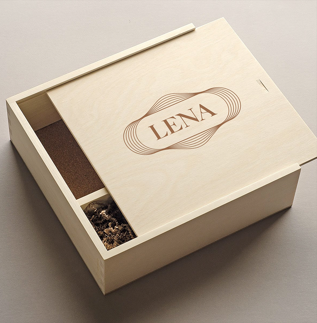 A wooden box with compartments, one containing brown packing material and another with decorative shredded paper, with the name "LENA" printed on the top.