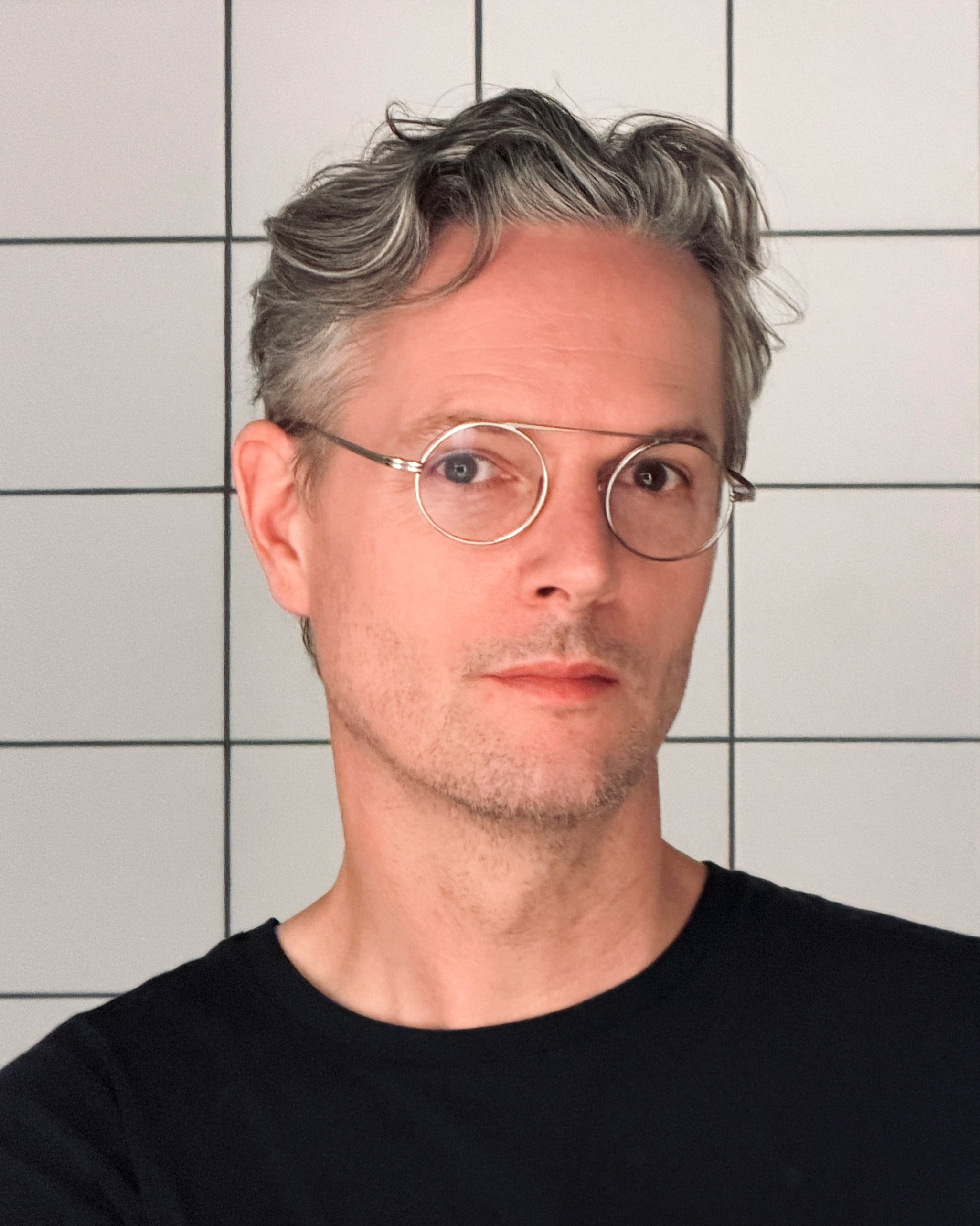 Close-up of a young man with wavy, grayish hair, wearing glasses and a black shirt, standing in front of a tiled wall.