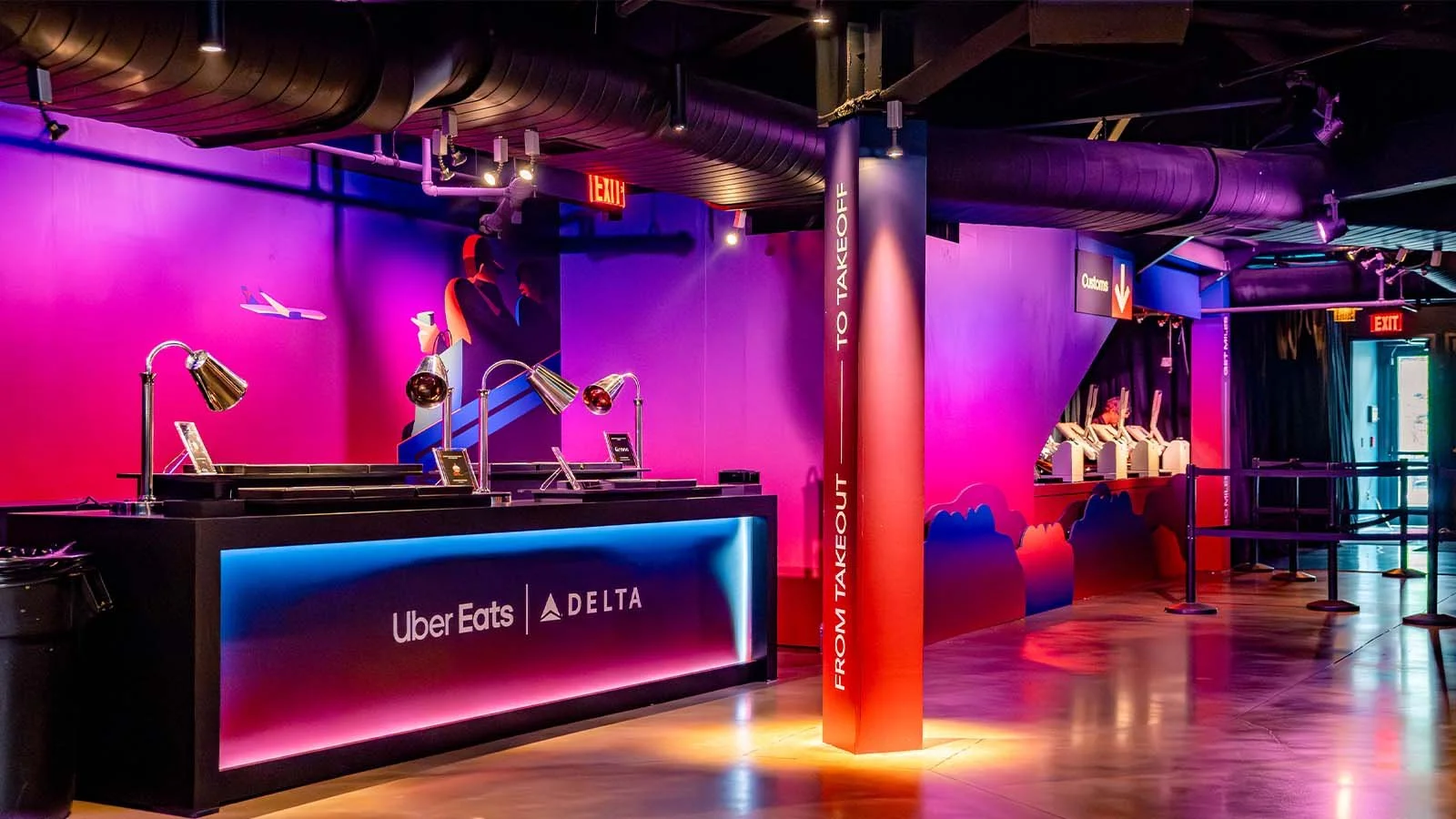 Interior of a brightly lit food pickup area with a counter displaying Uber Eats and Delta logos, pink and purple lighting, a sign indicating "From Takeout to Takeoff," and a line of computer stations with people at them in the background.