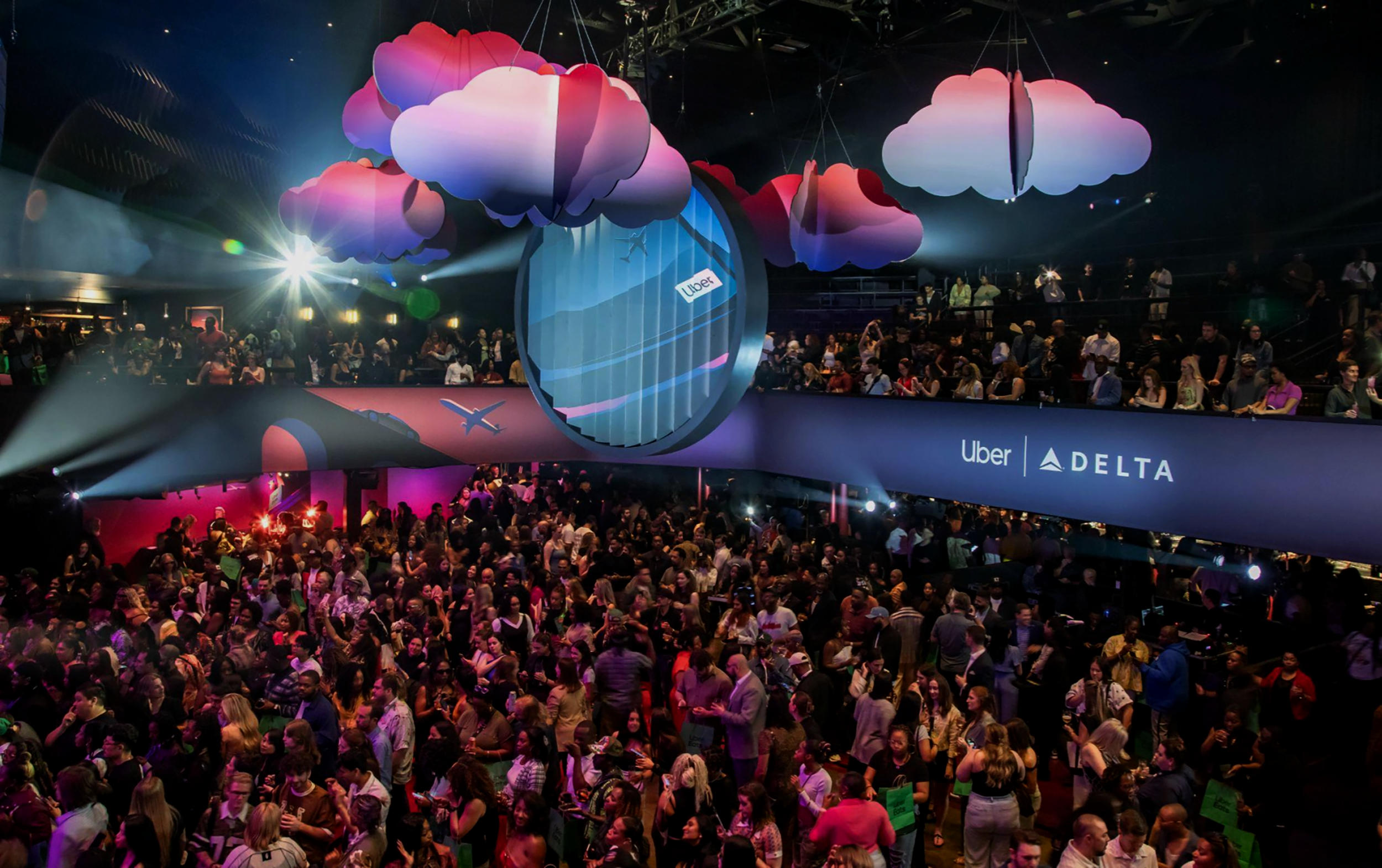 A large crowd of people gathered at an indoor event with colorful hanging cloud-shaped decorations and a sign displaying Uber and Delta logos.