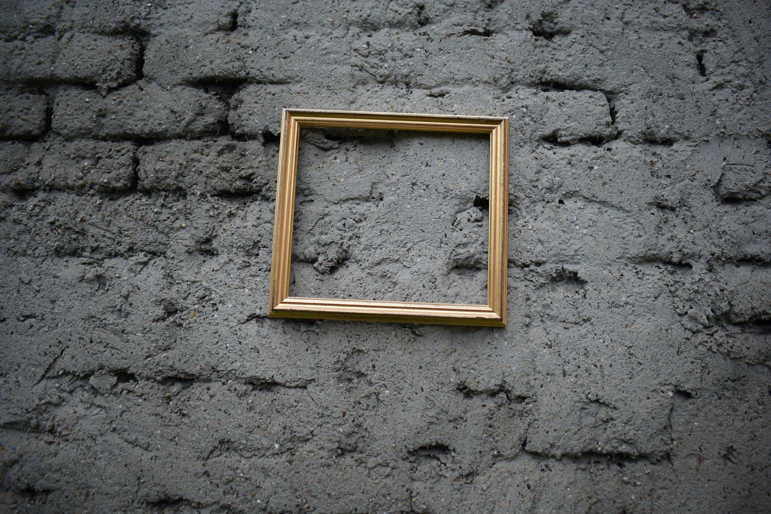An empty gold-colored picture frame resting on a rough gray brick wall. Framing and display services for fine art, contemporary art, sculpture and mixed media. Learn how to display your art.