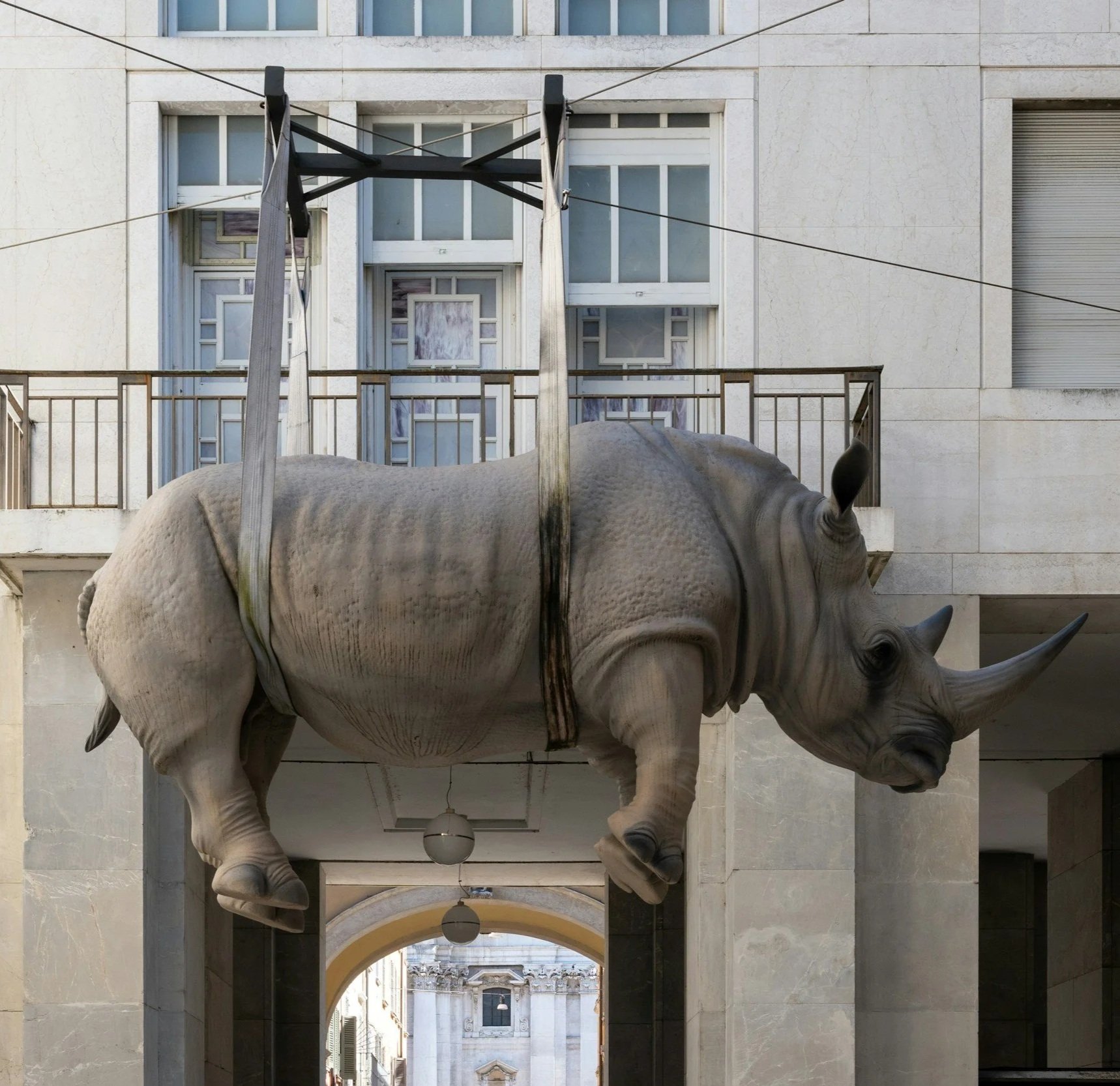 Street view showing a large rhinoceros sculpture attached to a building facade, with a balcony and architectural details in the background.