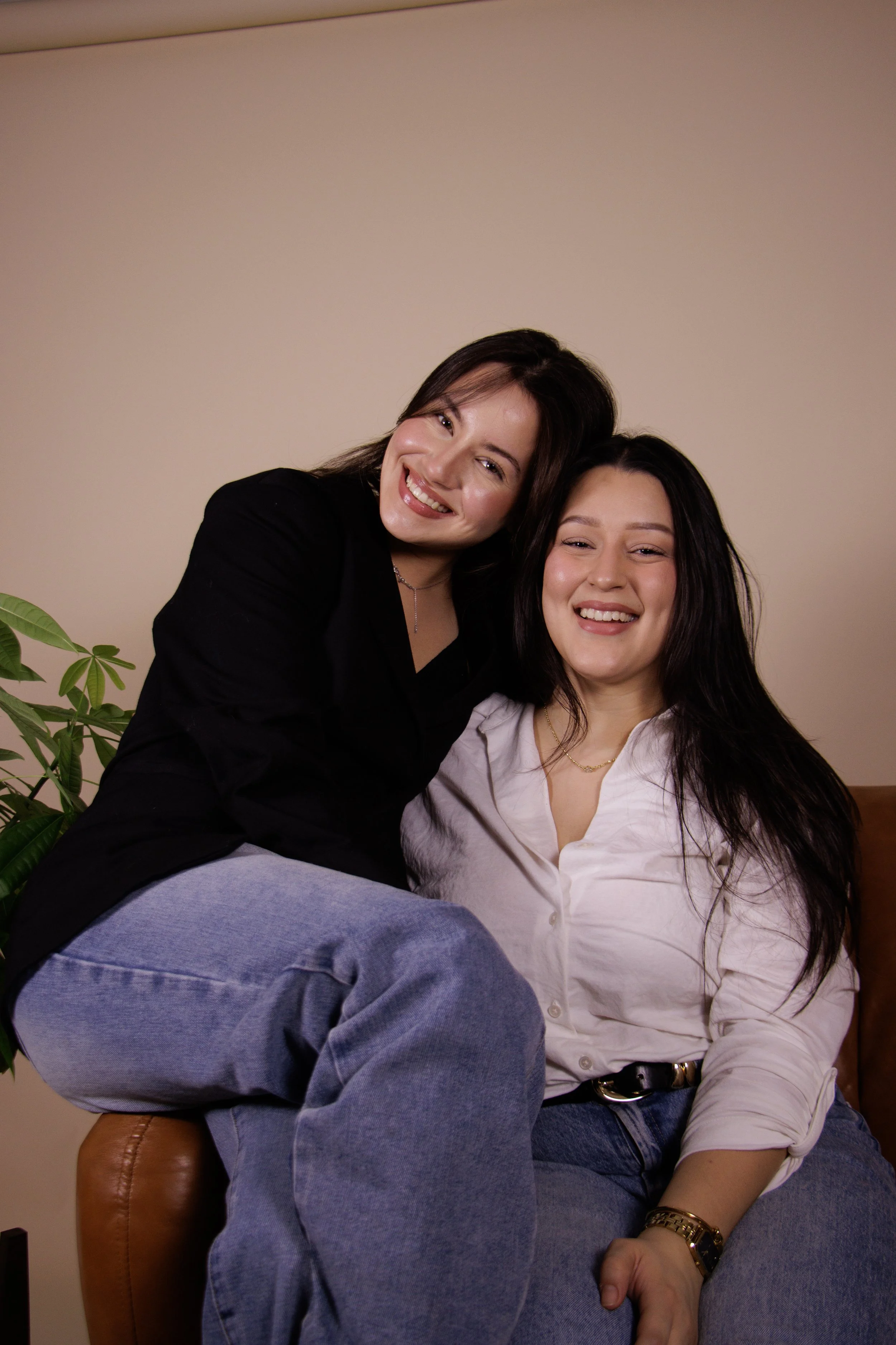 Two women sitting close together on a brown leather chair, smiling and looking at the camera. One woman with dark hair wearing a white shirt, the other with brown hair wearing a black jacket. A potted plant with green leaves is visible on the left side against a plain beige wall.