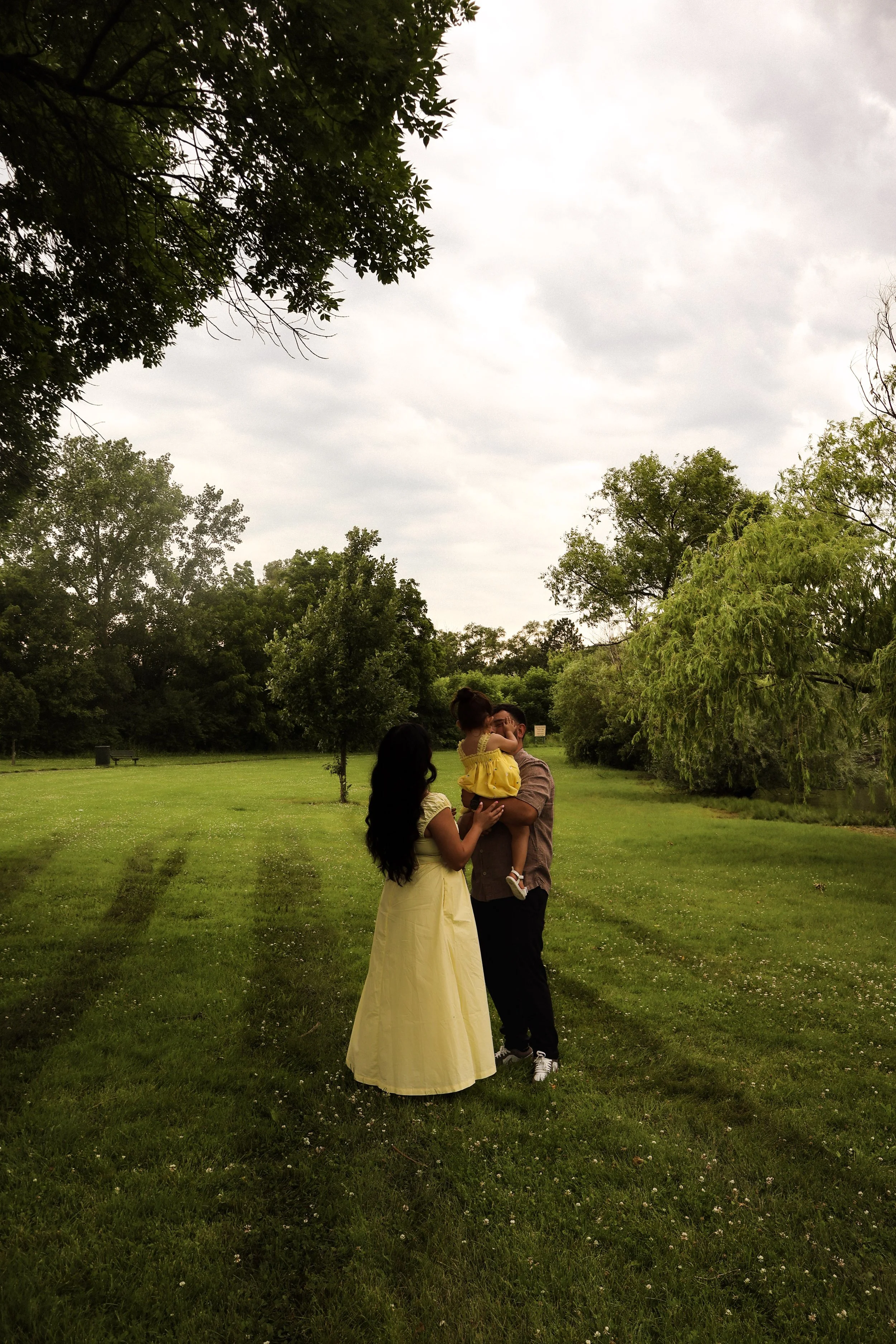 A family of three standing in a park, with a man holding a young girl in a yellow dress, and a woman in a long yellow dress. They are surrounded by green trees and grass, and the sky is cloudy.