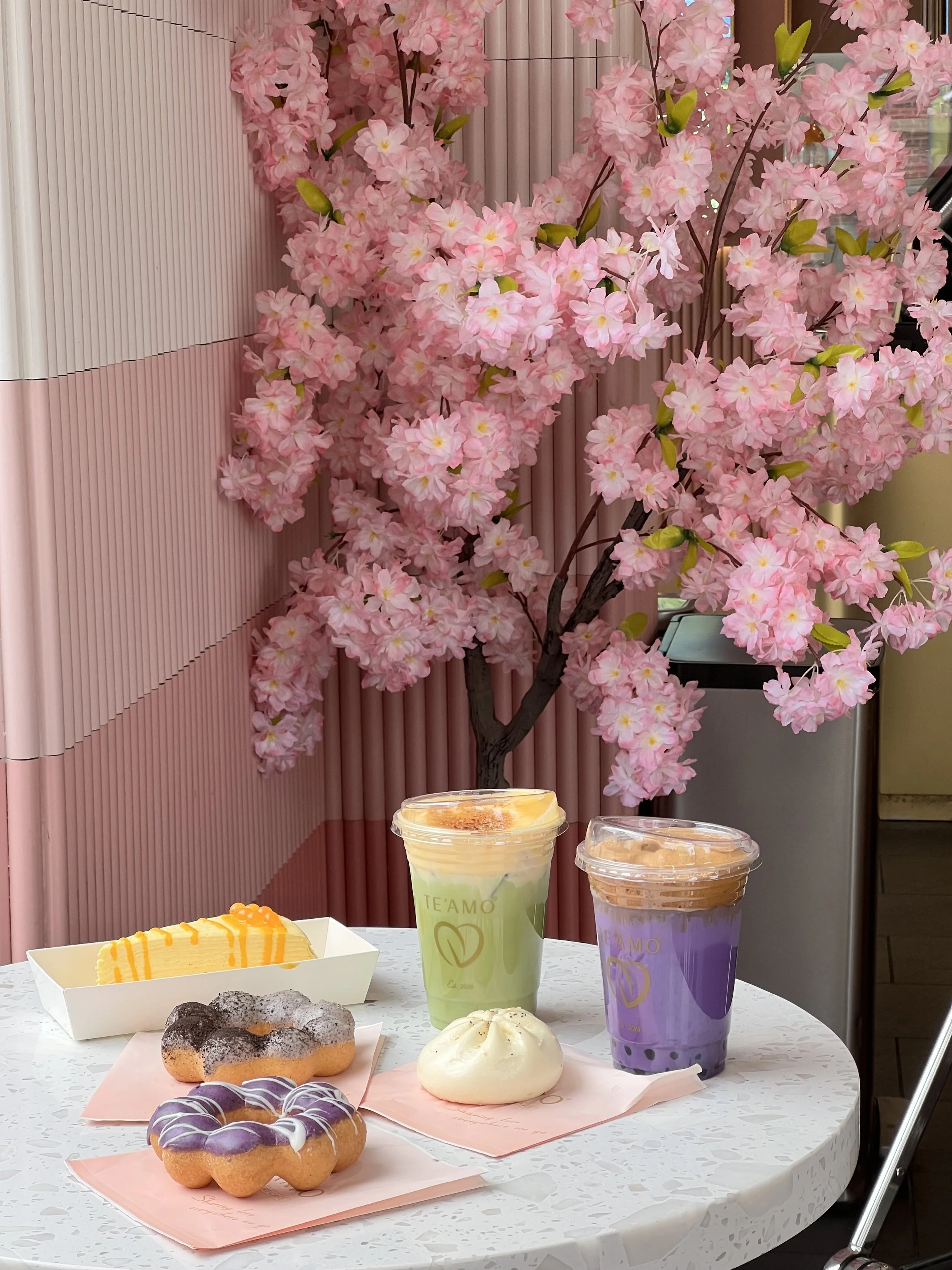 Pink artificial cherry blossom tree behind a table with assorted desserts and drinks including donuts, a green beverage, a purple beverage, and a piece of cake, set against a pink vertical slat background.