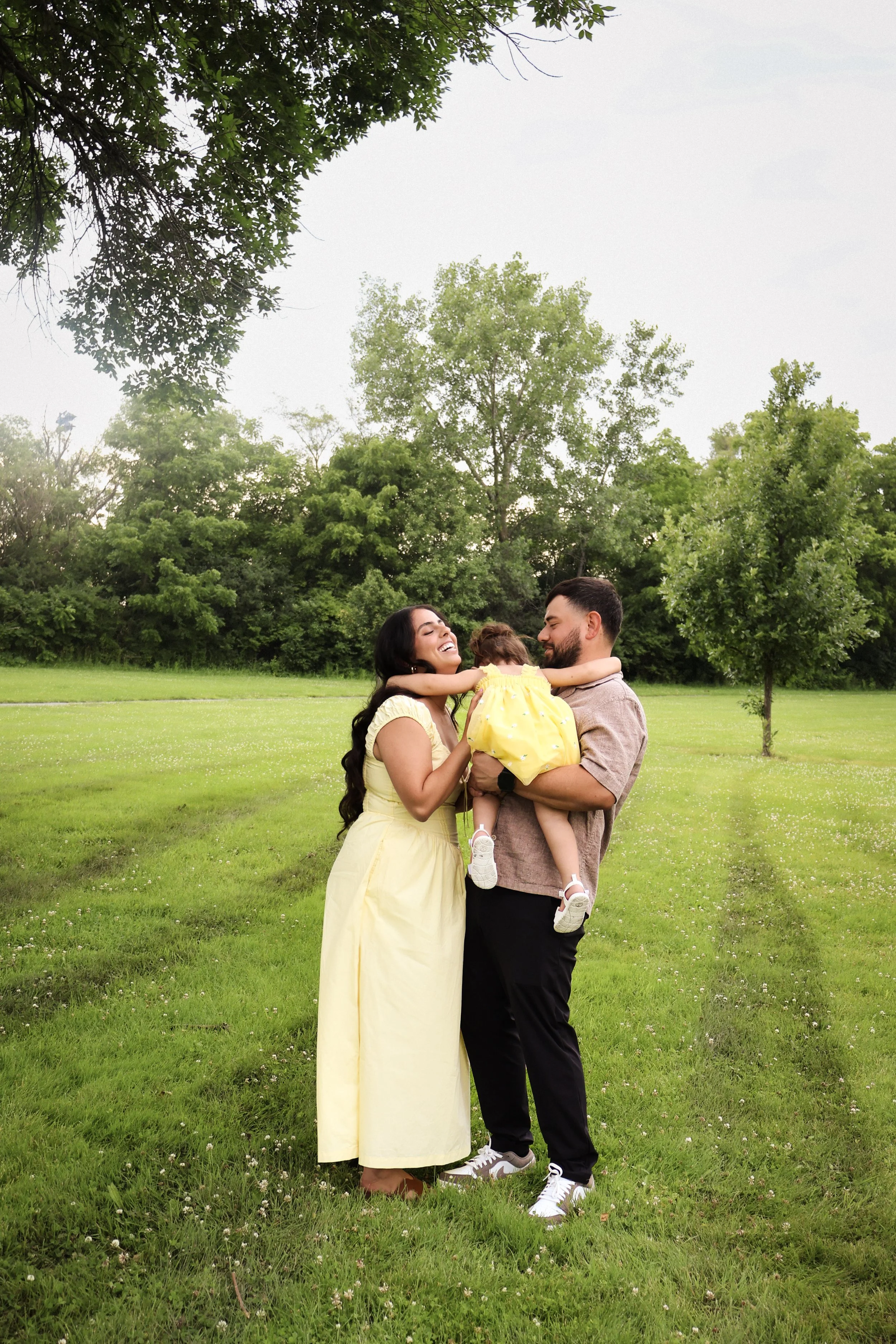 A family of three enjoying time together in a park during daytime. The woman, dressed in a yellow dress, is smiling while holding a young child wearing a yellow dress. The man, wearing a gray shirt and black pants, is smiling and holding the child. T