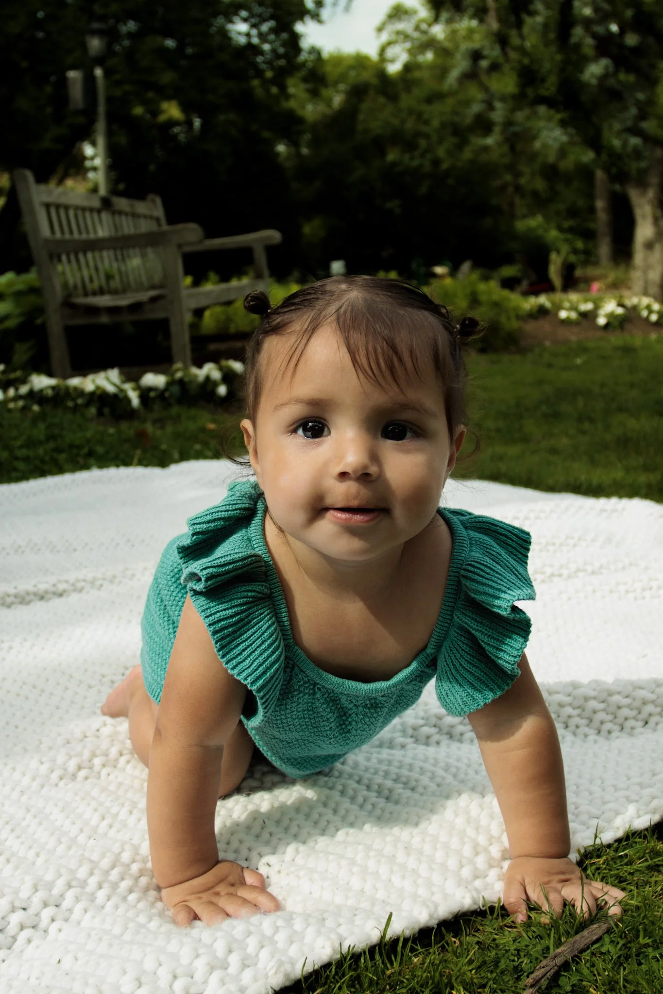A baby crawling on a white blanket outdoors in a garden with green grass, flowers, trees, and a wooden bench in the background.