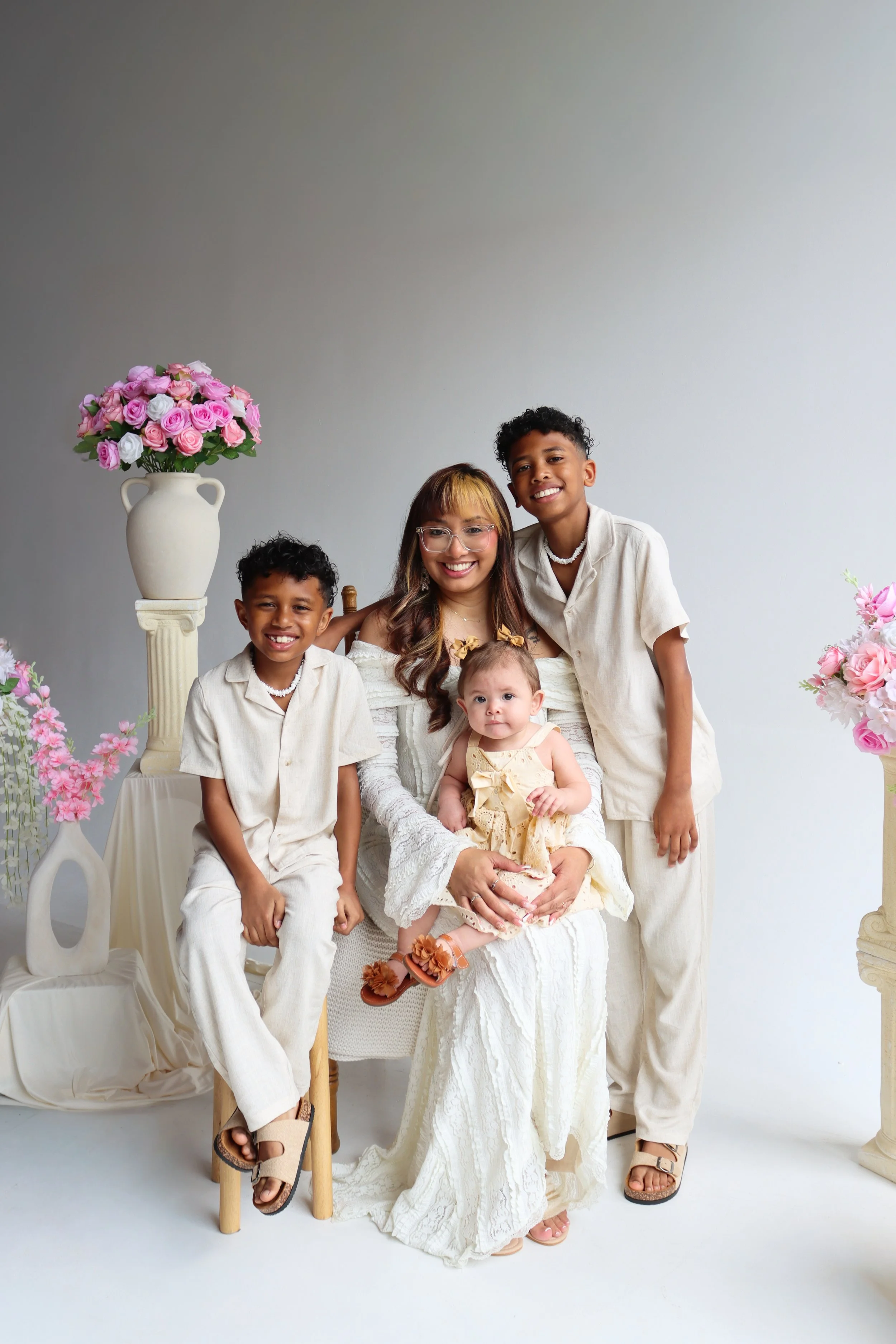 A woman with three children sitting and standing in front of a white background, surrounded by large floral arrangements of pink and white roses in vases