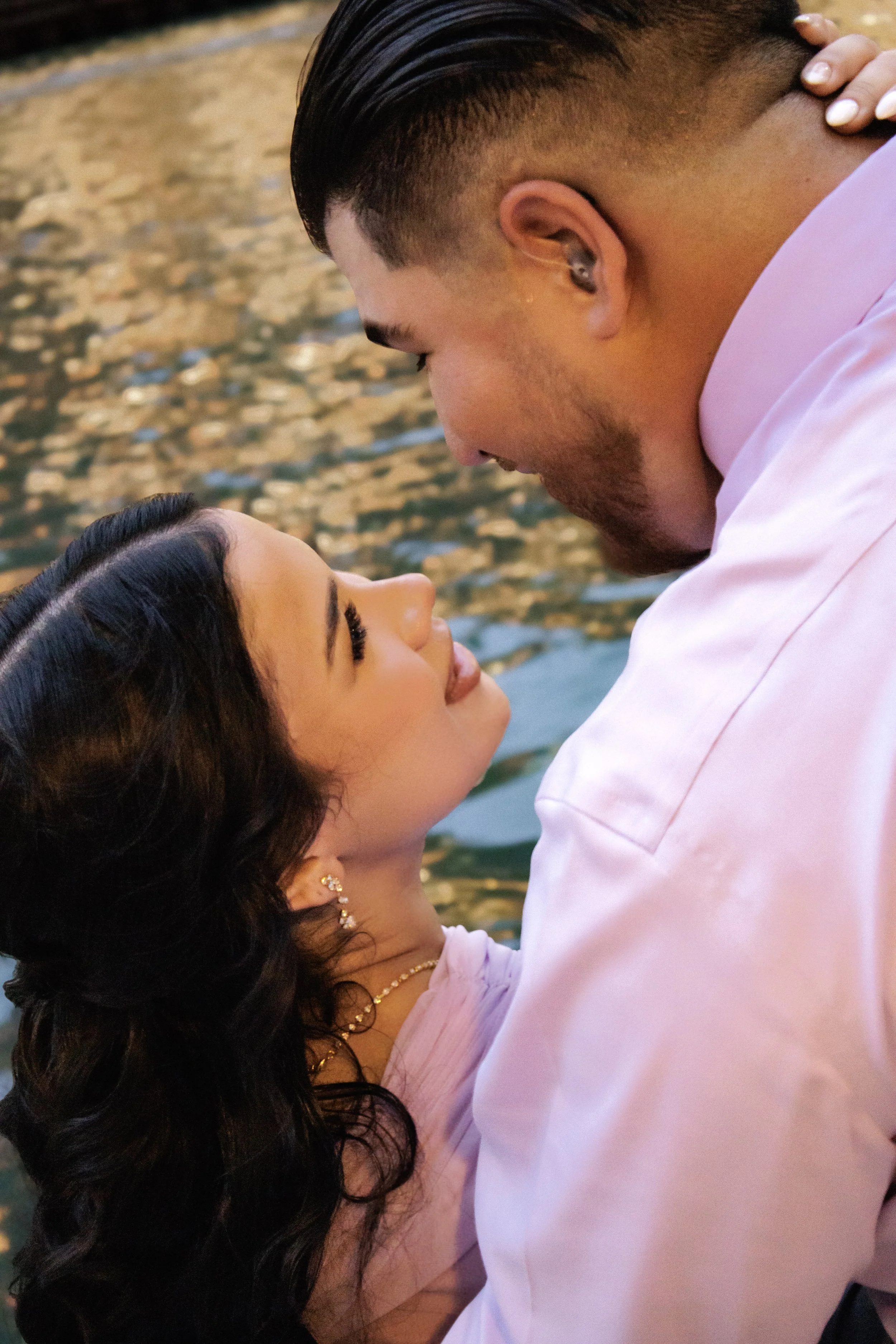 A man and woman are close to each other, gazing into each other's eyes near a body of water with gentle ripples.