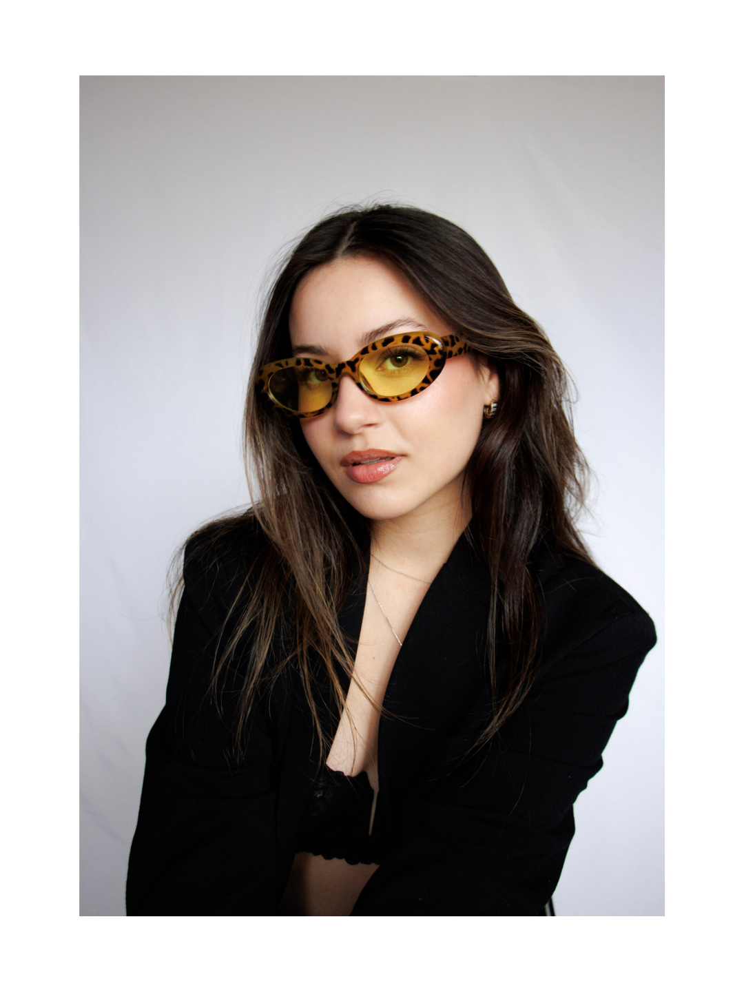 A woman with long dark hair wearing leopard-print sunglasses with yellow lenses, black blazer, and a delicate necklace, posing against a plain white background.