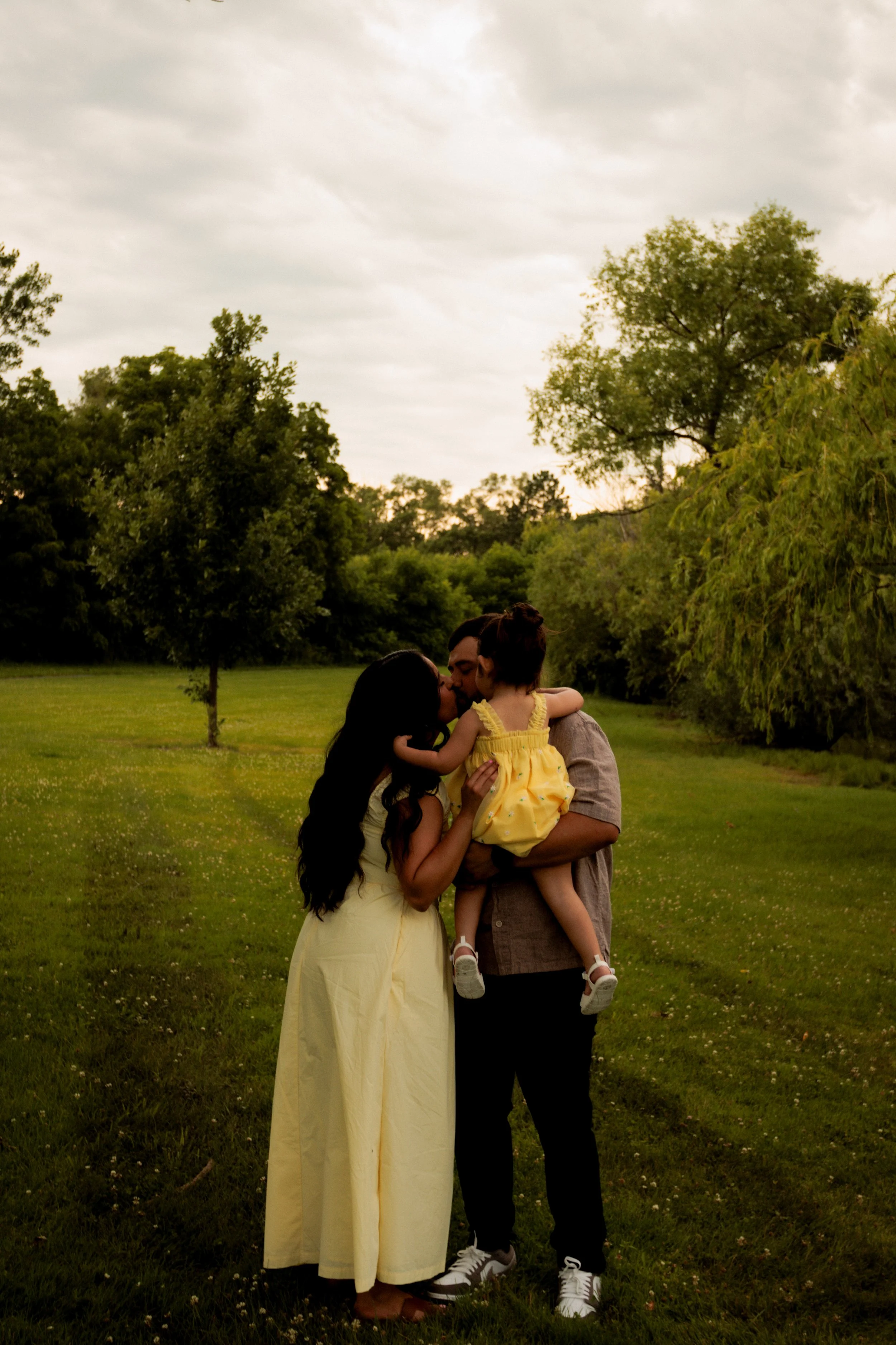 A family of three standing in a grassy park, with daughter being held by father and mother kissing daughter, all surrounded by trees under cloudy sky.