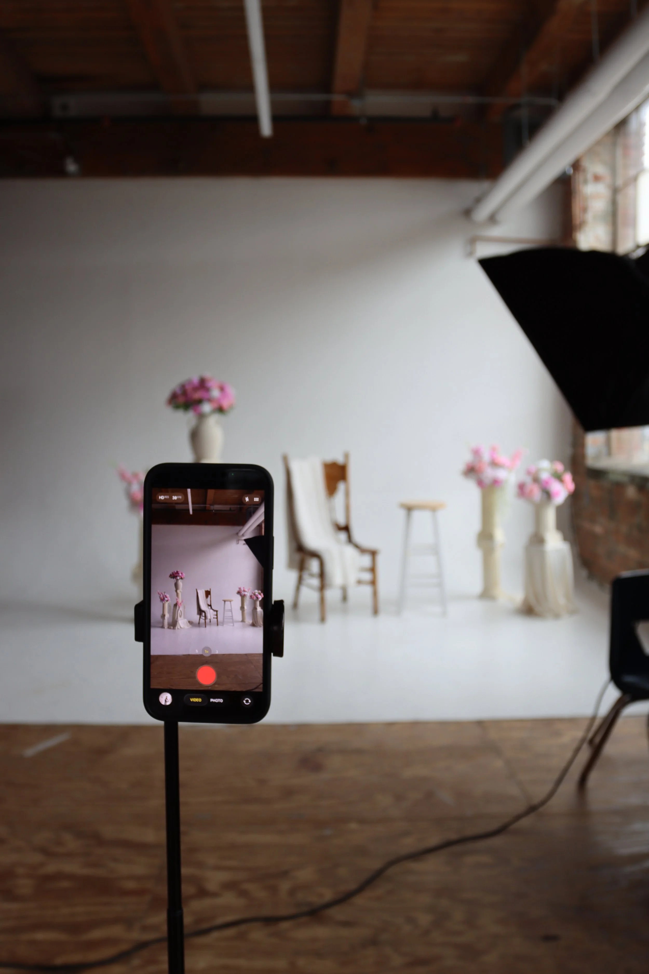 A photo of a web photo shoot setup with a smartphone on a tripod recording a background setup of chairs and flower arrangements in vases, with lighting equipment and a partially visible window brick wall to the right.