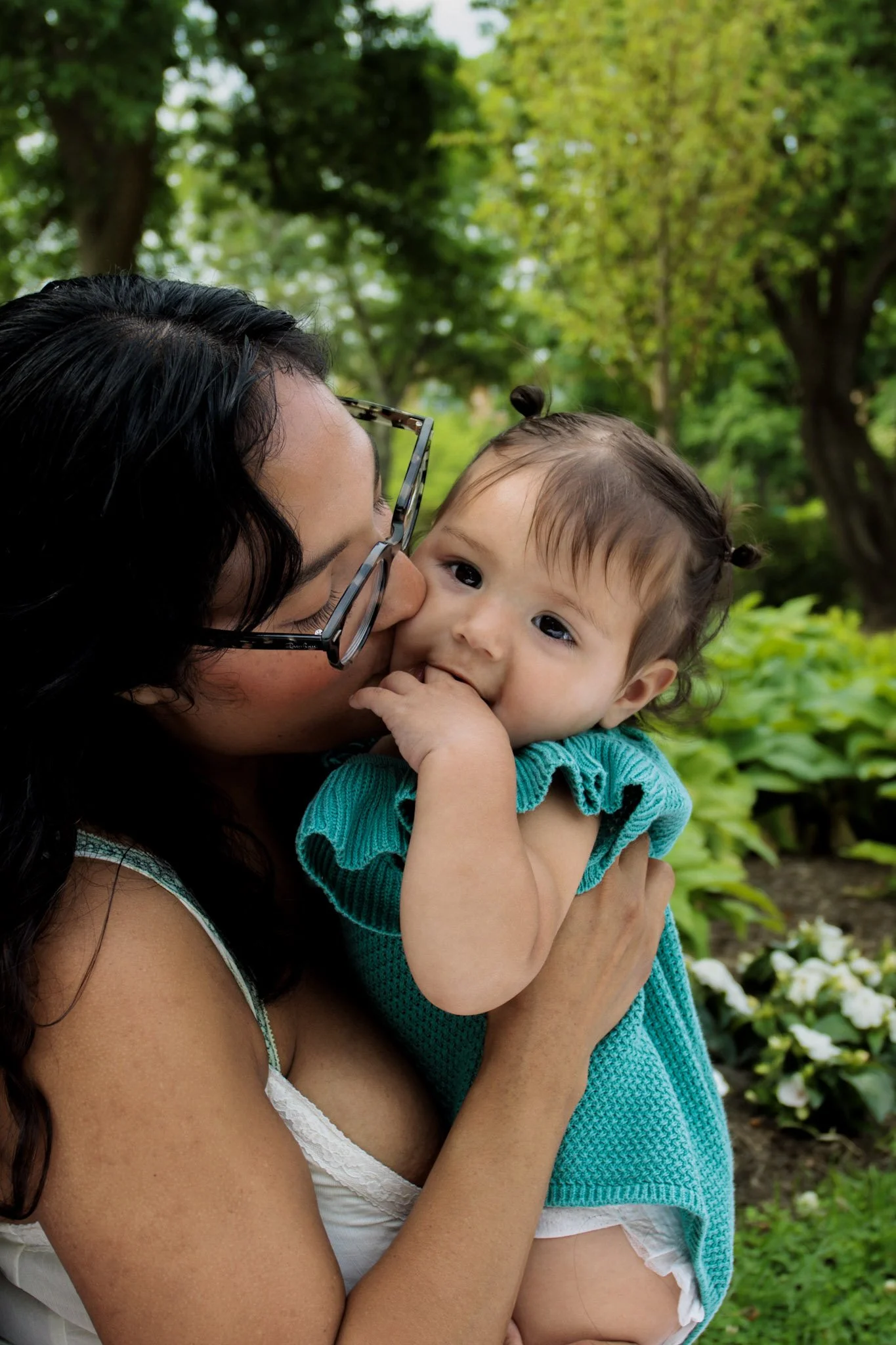 A woman with dark hair and glasses holding a young girl with pigtails, both outdoors in a park with green trees and flowers in the background. The woman is kissing the girl on the cheek, and the girl is smiling with her fingers in her mouth.