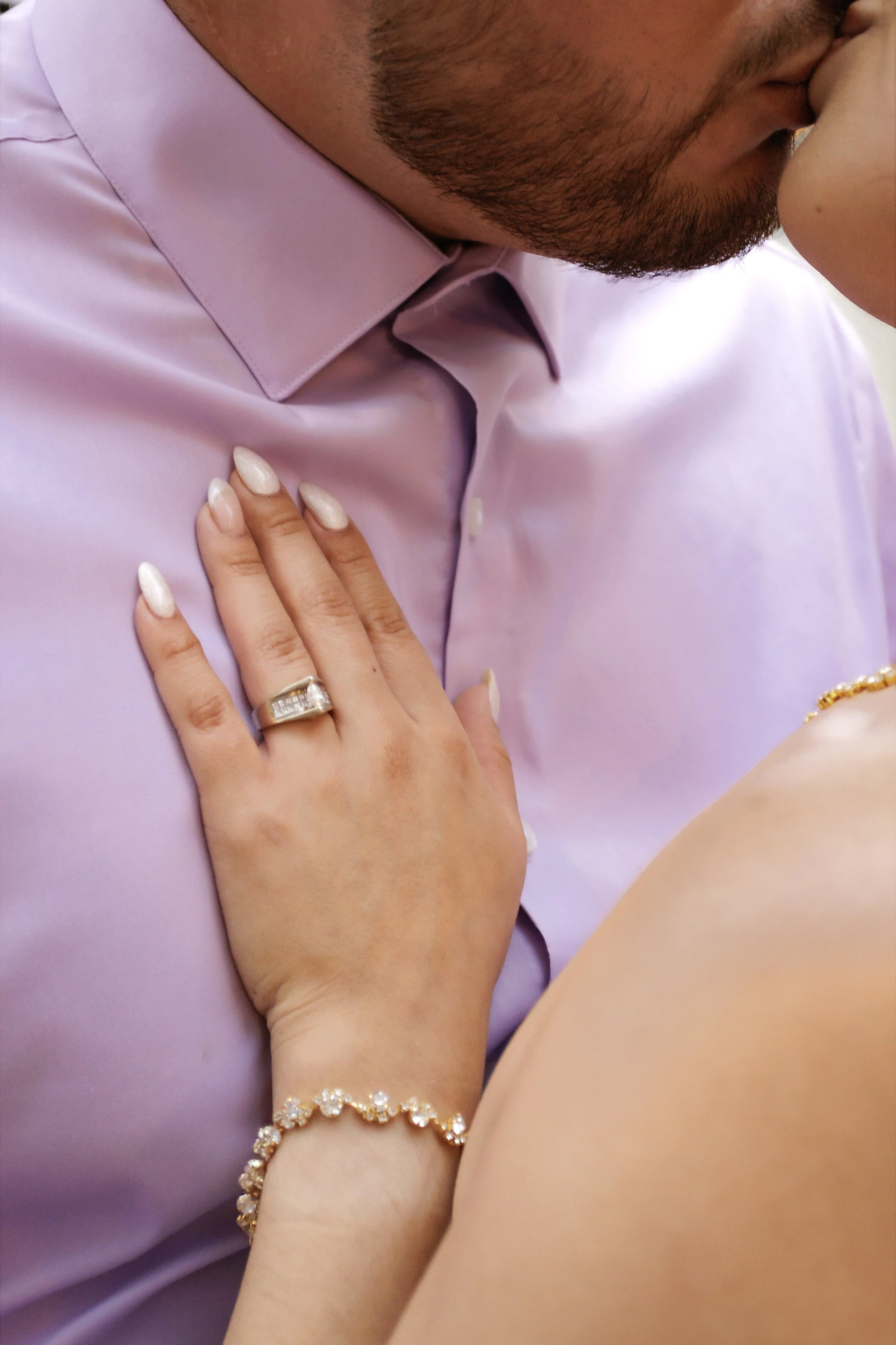 A person wearing a lavender-colored shirt and a woman with a bracelet and ring is holding and leaning against a man in a lavender shirt.