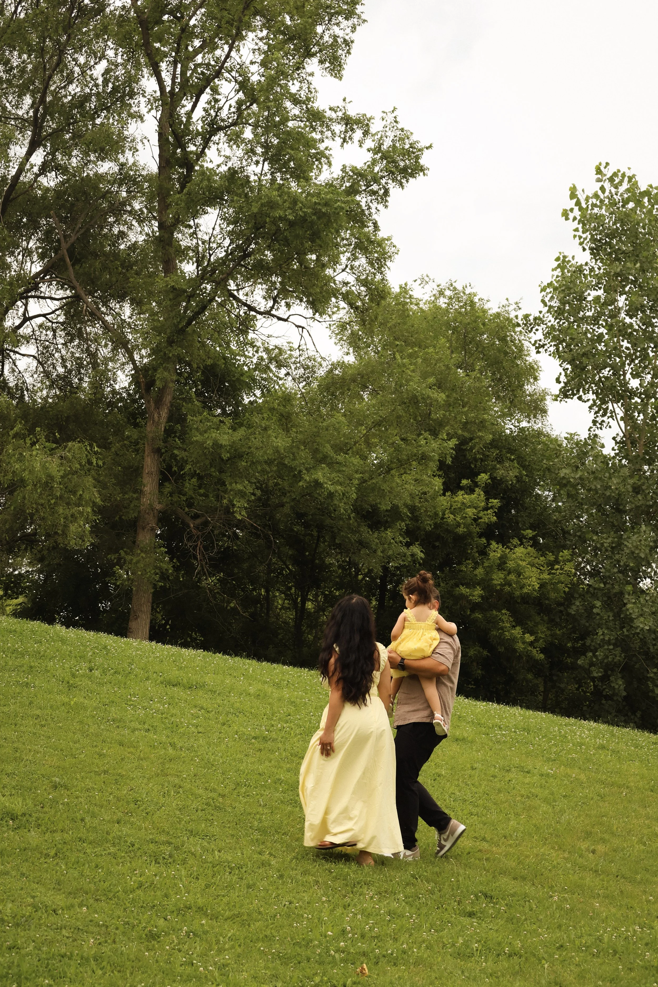 A woman walking on grass with a man carrying a child while holding hands with another woman, all outdoors with trees in the background.