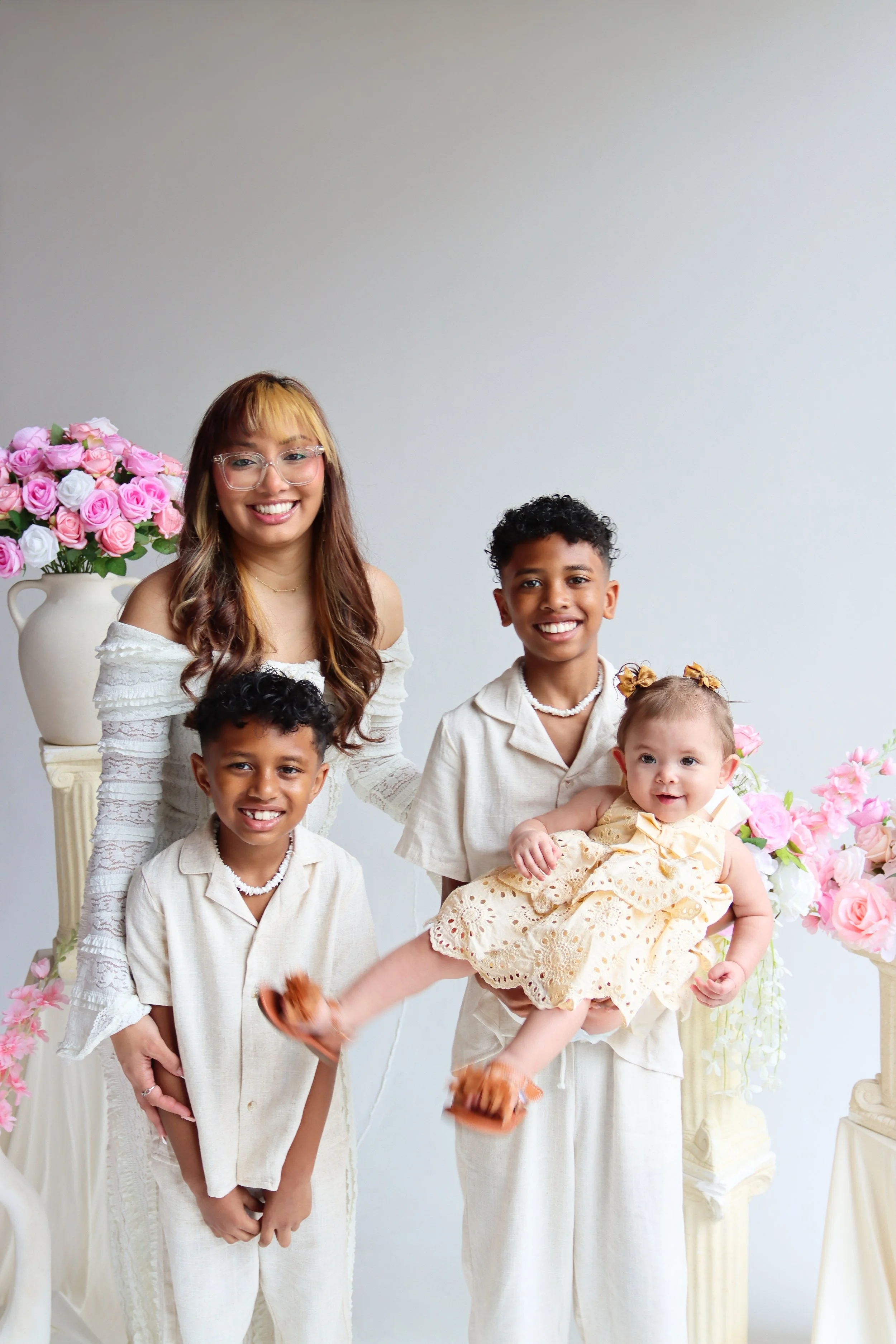 A woman with glasses and long hair, smiling, standing next to four children, three boys and a girl, surrounded by pink and white roses, in a light-colored room with a plain wall background.