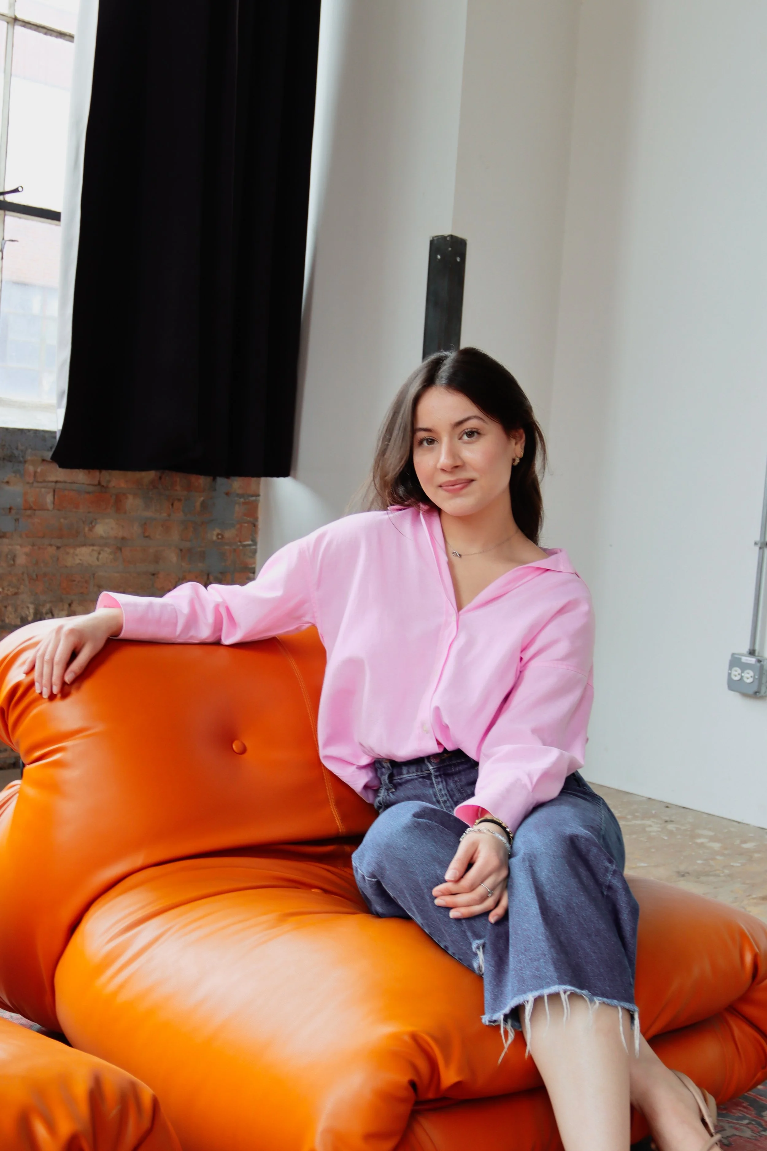A young woman with brown hair and light skin, wearing a pink hoodie and blue jeans, sitting on an orange leather couch in a room with white walls, black curtains, and a brick wall visible through the window.