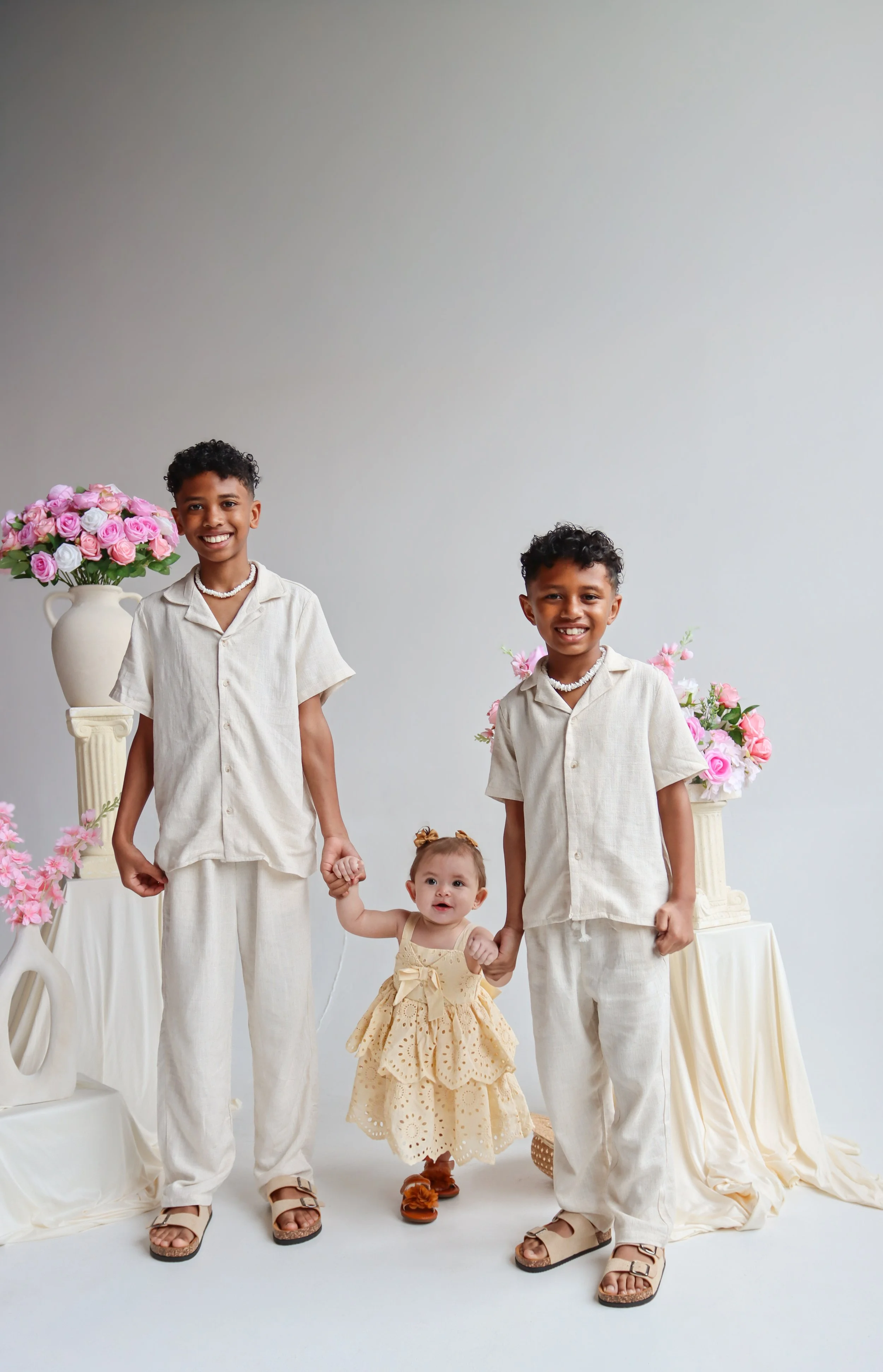 A group of four children standing in front of a plain white backdrop, holding hands. They are dressed in light-colored loose-fitting clothing. Two of the children are boys, one is a girl, and the youngest child is a baby girl. There are pink and whit