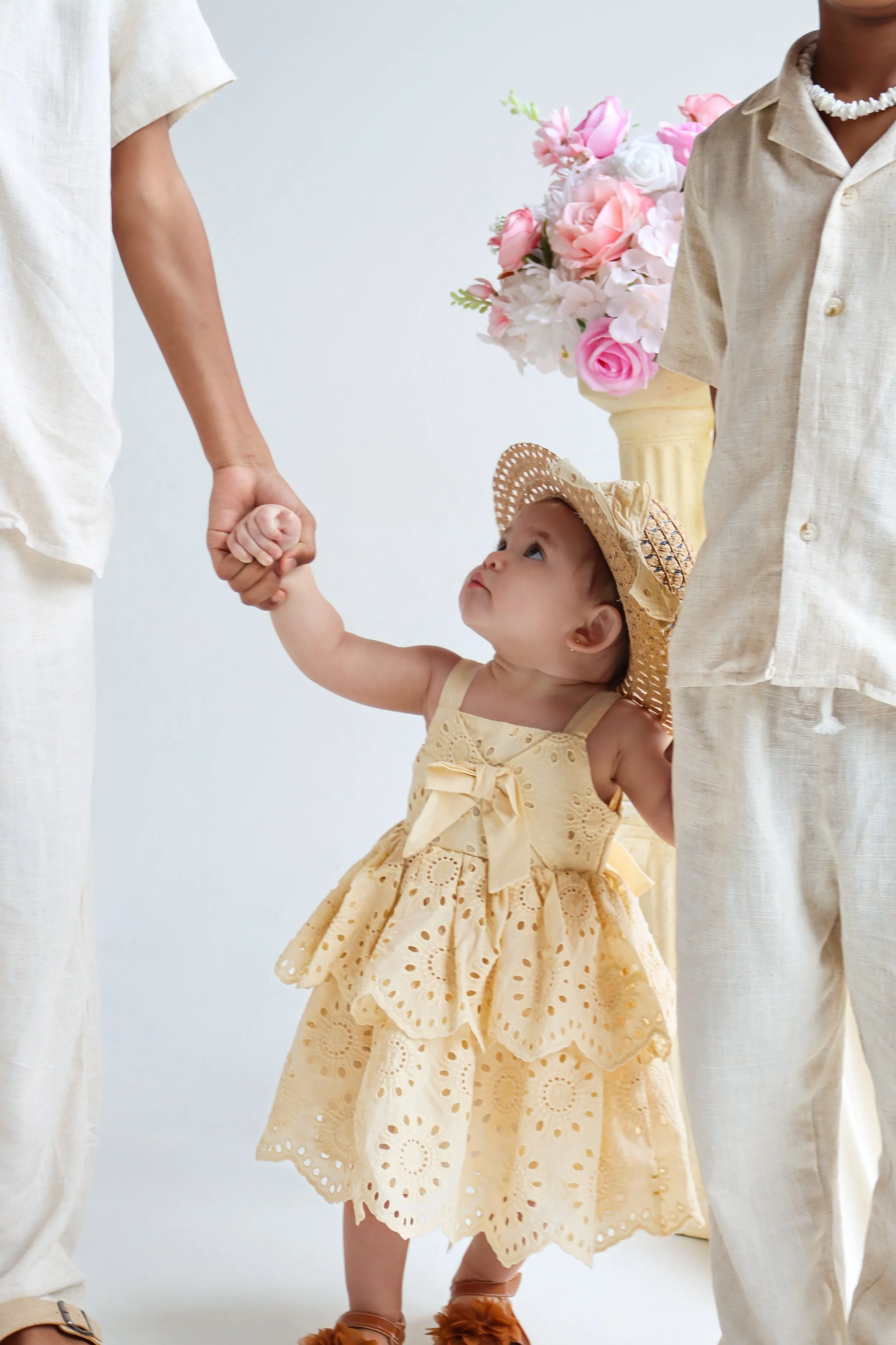 A young girl wearing a yellow eyelet dress and a straw hat, holding hands with two adults dressed in light-colored clothing, standing in front of a vase of pink and white flowers.