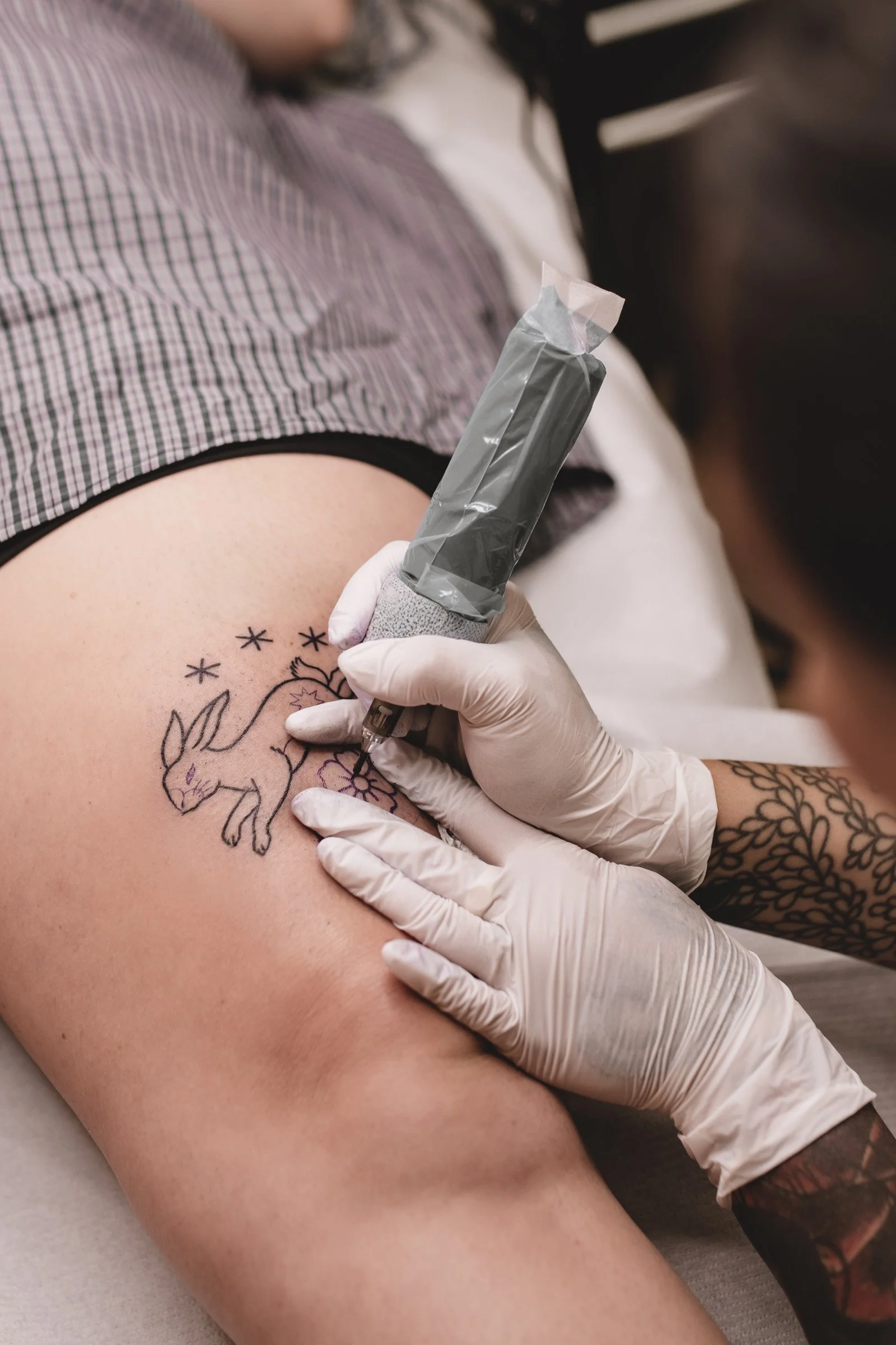A person is getting a tattoo of a unicorn on their thigh by a tattoo artist wearing gloves.