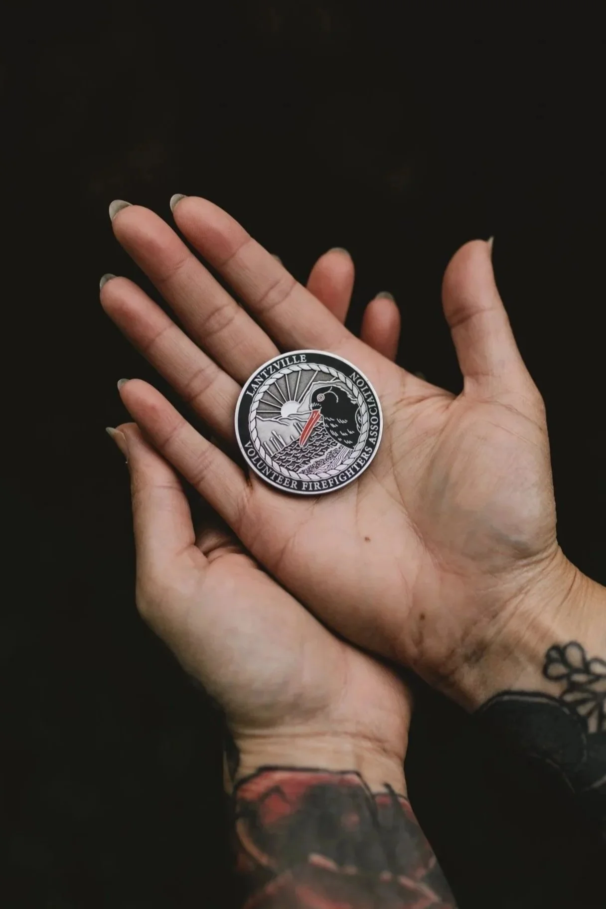 A hand holding a round patch with a black bird, a sun, and the words 'LANTZVILLE VOLUNTEER FIREFIGHTERS ASSOCIATION' on it.