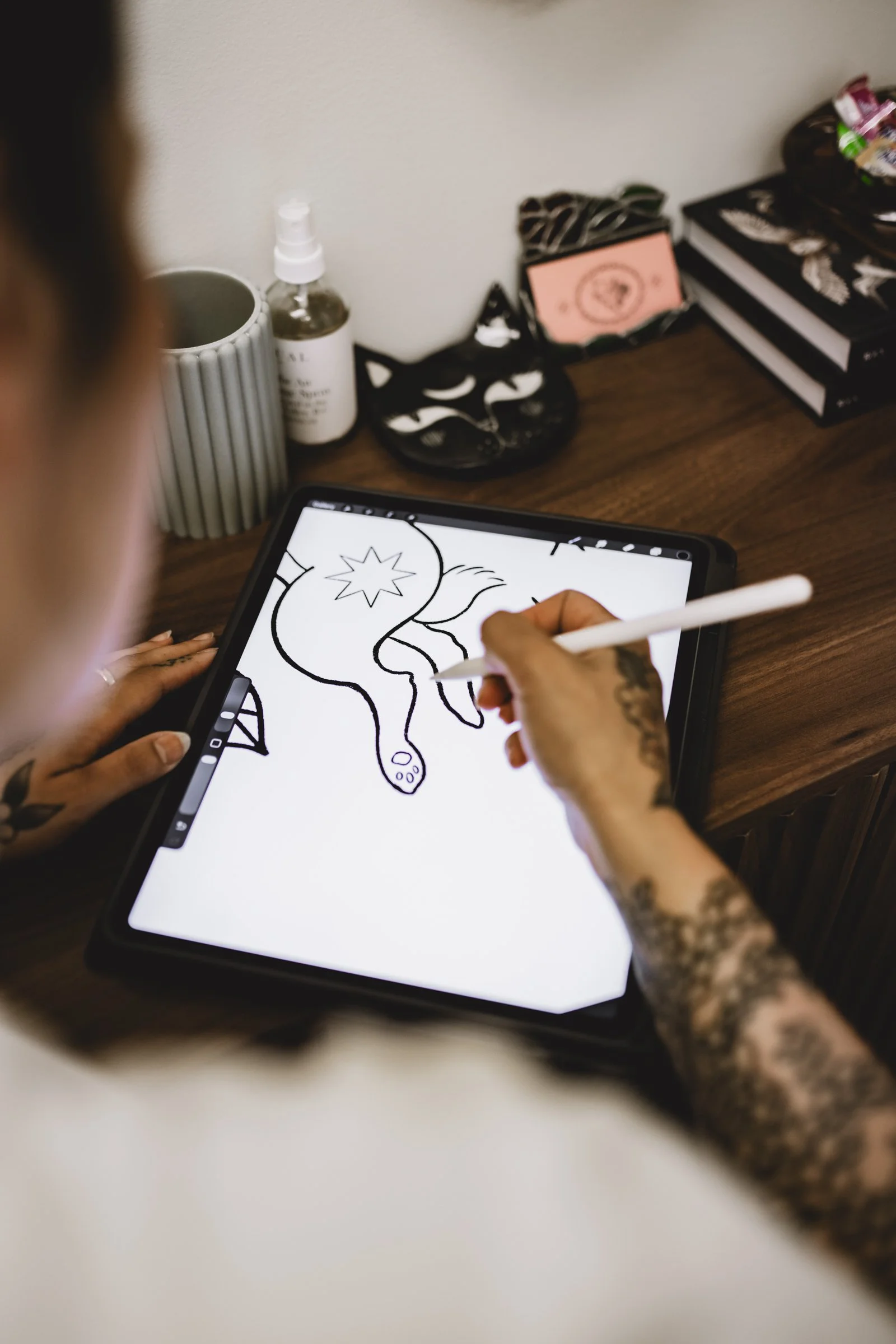 Person with tattoos drawing a unicorn on a digital tablet using a stylus, with various books and decorative items on a wooden desk.