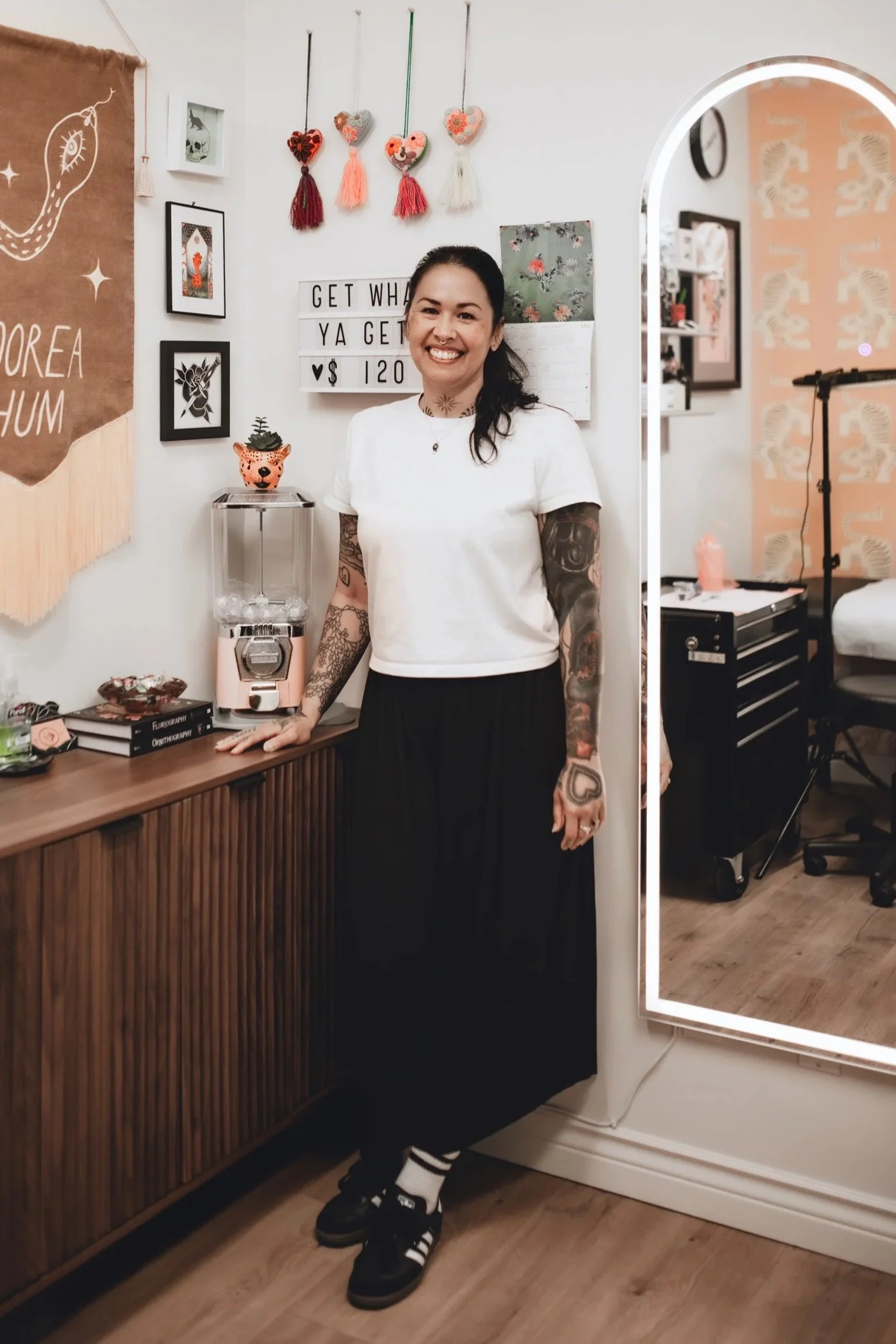 A woman with tattoos on her arms, smiling and standing indoors near a wooden desk and a mirror. She is wearing a white T-shirt, black maxi skirt, and black sneakers with white stripes.