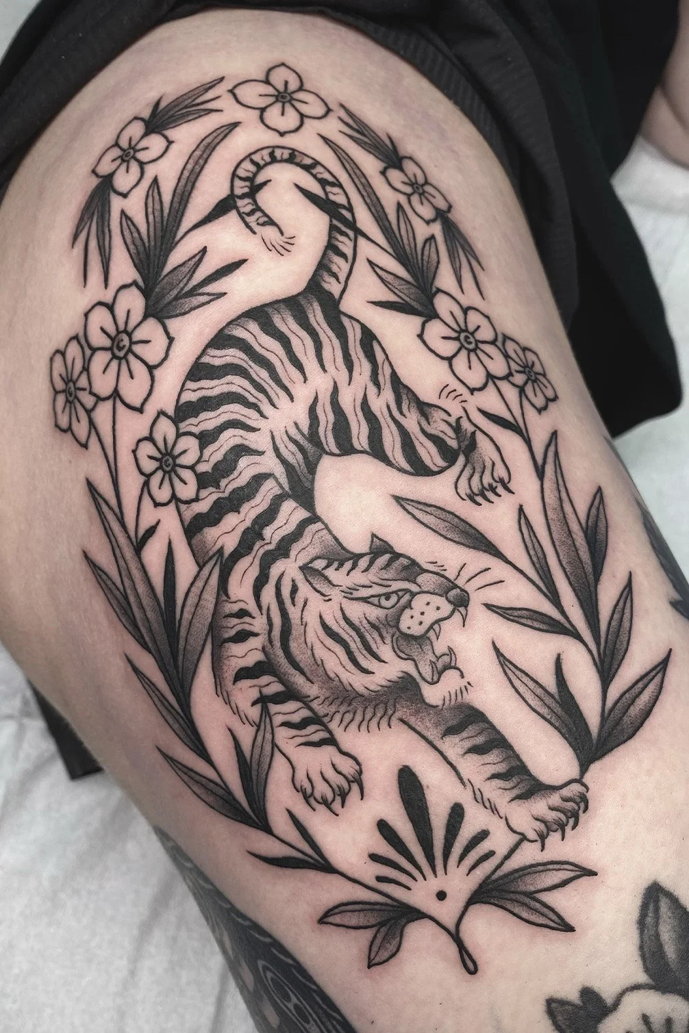 Black and gray tattoo of a tiger surrounded by flowers and leaves on a person's thigh.