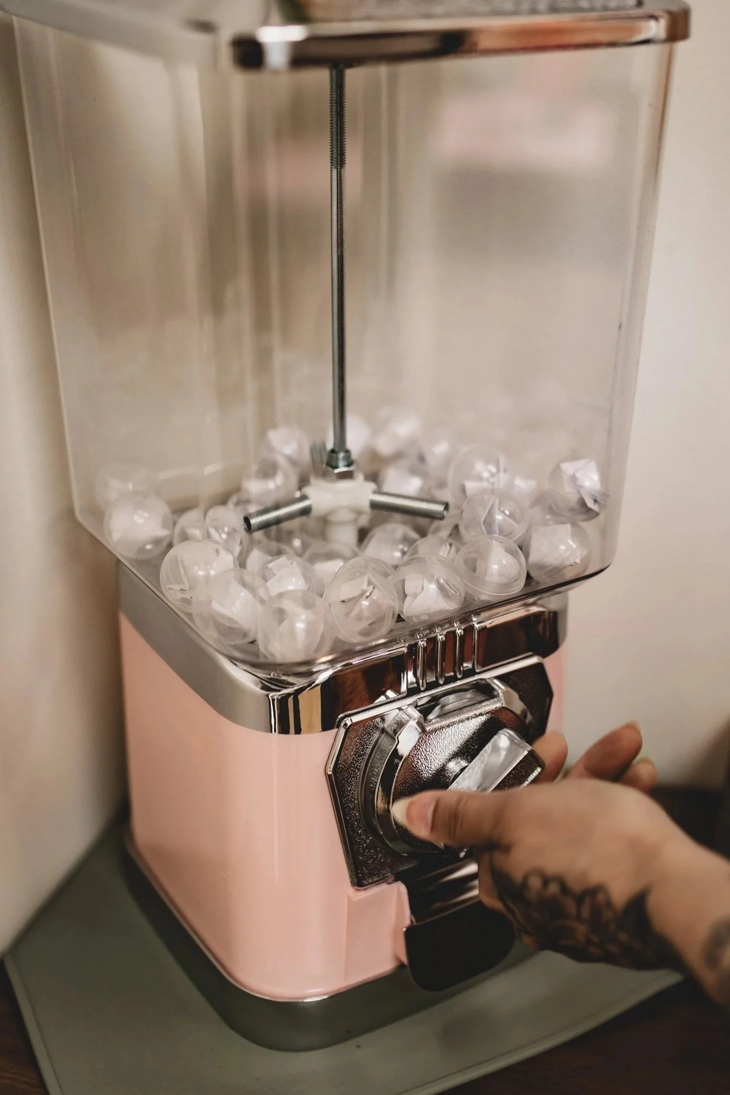 A person with a tattooed hand turning a dial on a pastel pink vintage-style espresso machine with a clear container filled with empty pods on top.