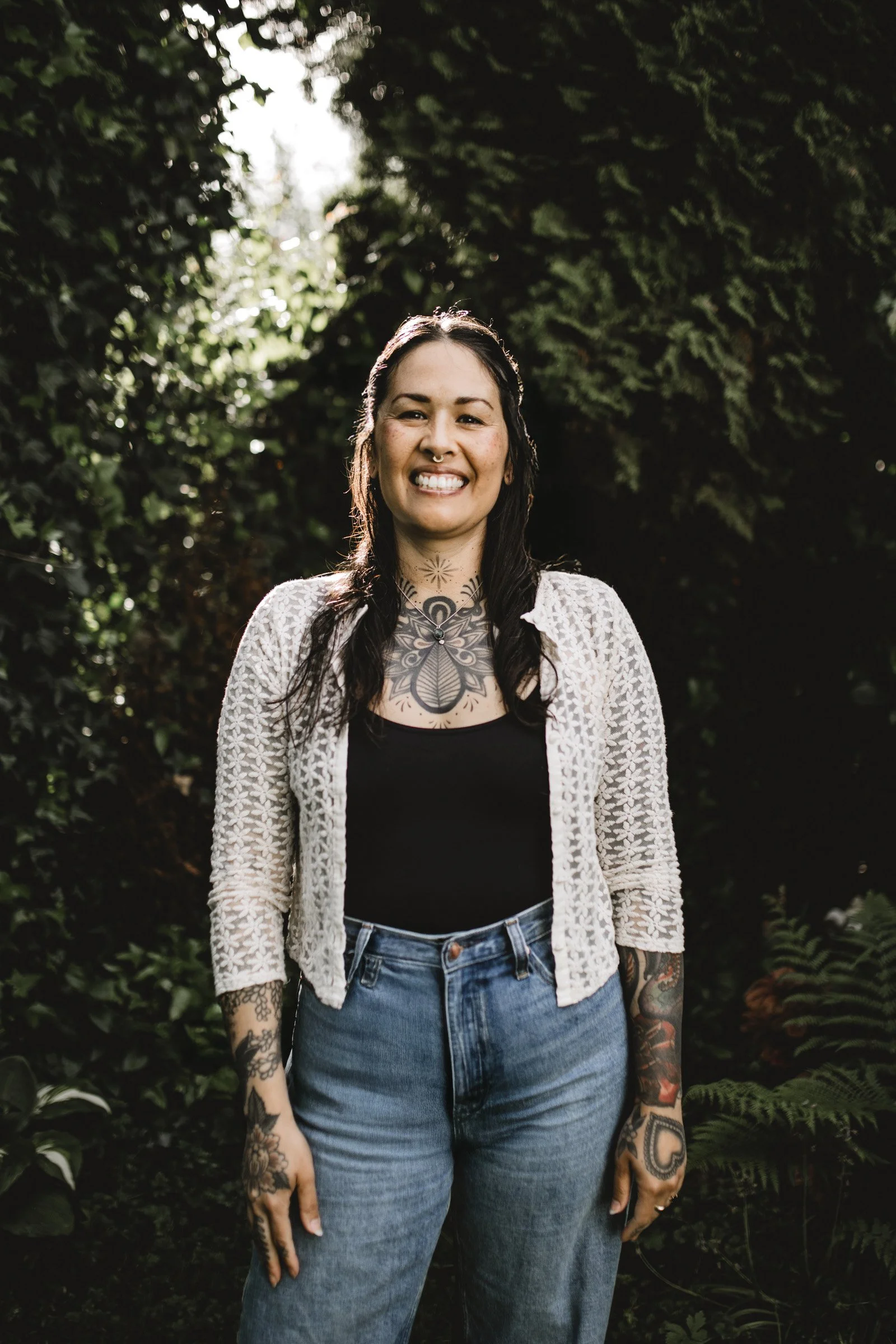 A smiling woman with tattoos on her arms and chest, wearing a black top, a white textured cardigan, and jeans, standing outdoors in front of green foliage.