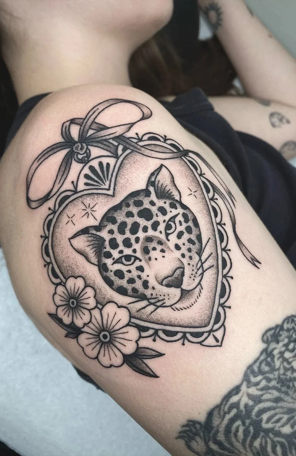 Black and gray tattoo of a leopard's face inside a heart-shaped frame decorated with flowers and a bow on top.