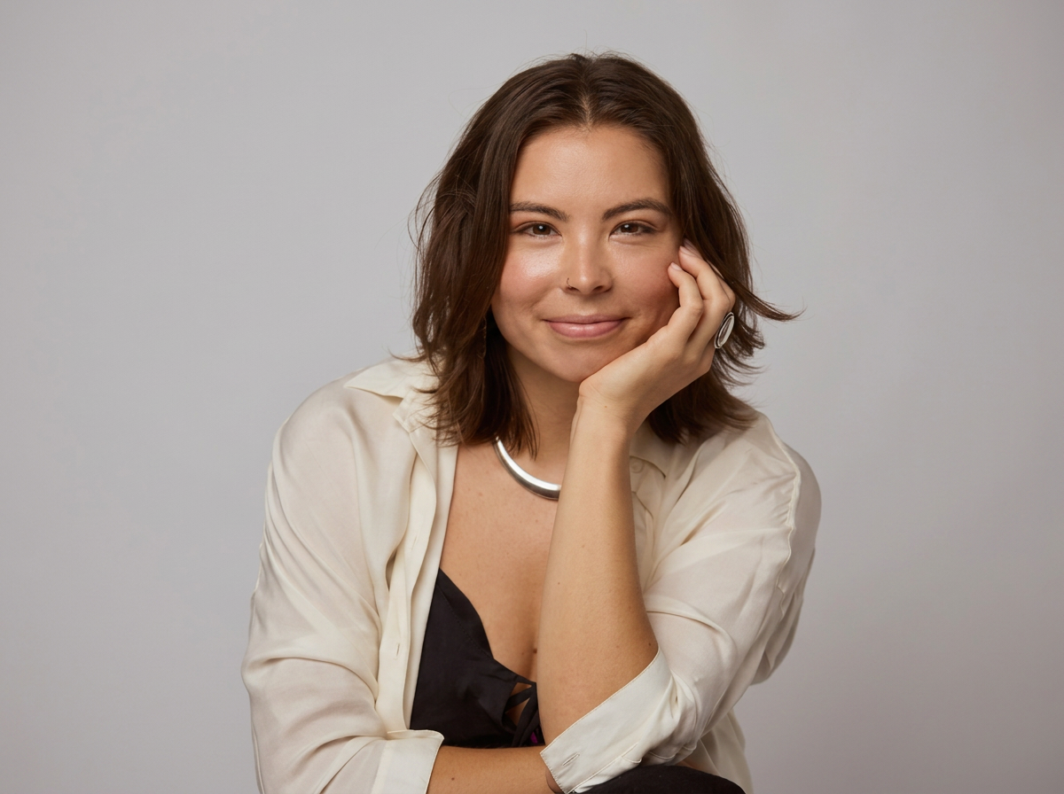 A young woman with shoulder-length brown hair, smiling gently, resting her face on her hand, wearing a cream-colored blouse and silver jewelry, against a plain light gray background.