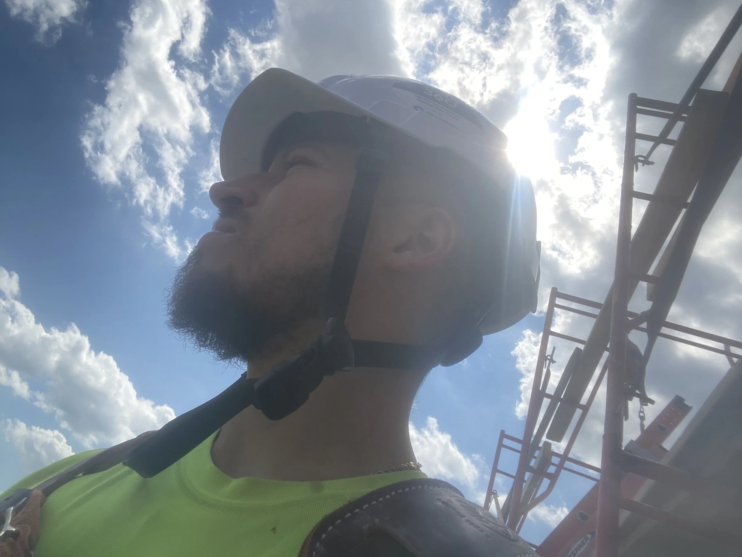 A person wearing a helmet and a bright green shirt, taking a selfie at an amusement park or fair with a roller coaster and blue sky with clouds in the background.