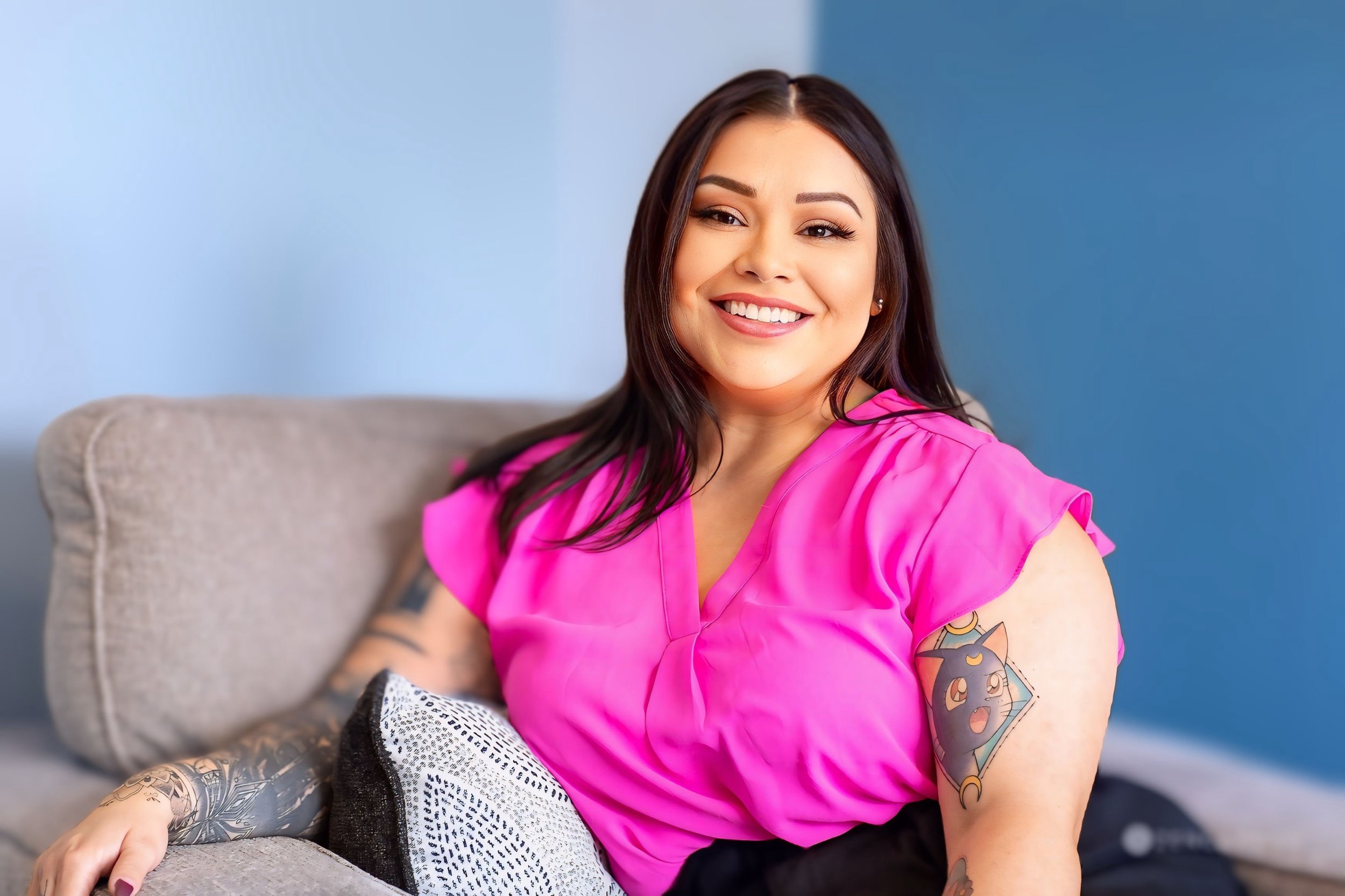 A woman with long dark hair, tattoos on her arm, wearing a bright pink top, sitting on a gray couch with a blue wall in the background, smiling at the camera.