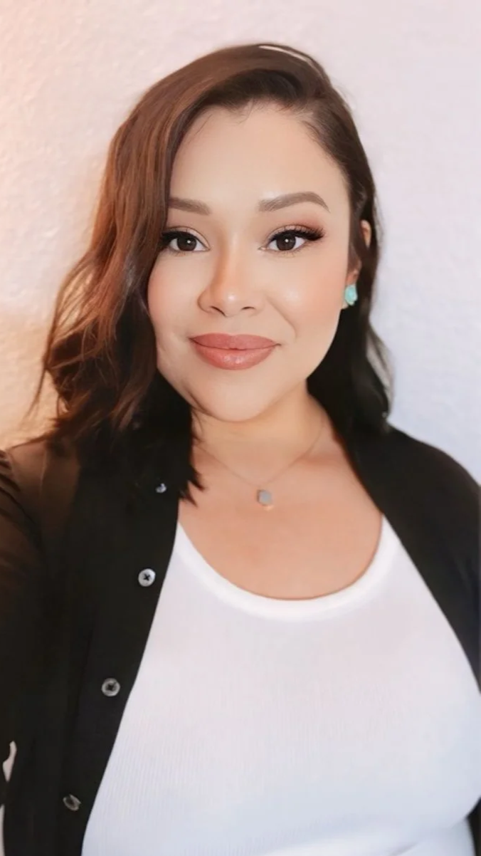 A woman with shoulder-length dark brown hair, wearing makeup, a white top, black jacket, and teal earrings, smiling at the camera against a neutral background.