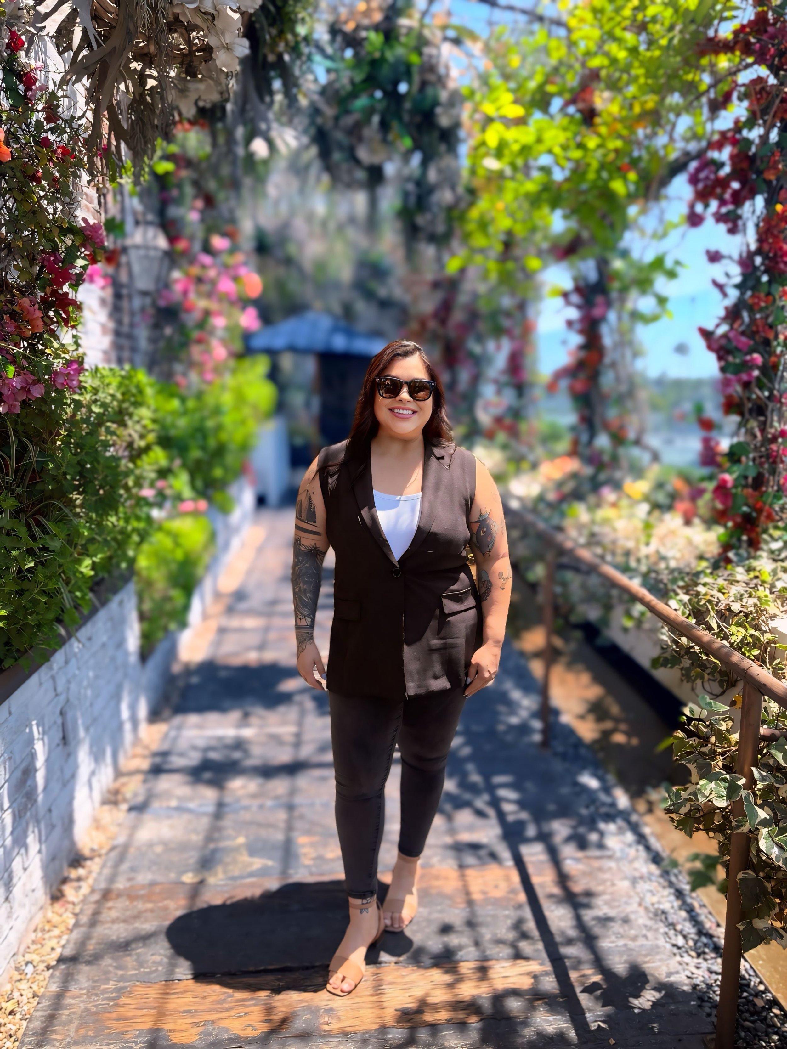 A woman with tattoos on her arms wearing sunglasses and a black sleeveless blazer over a white shirt, smiling while walking on a pathway surrounded by colorful flowers and green foliage on a sunny day.