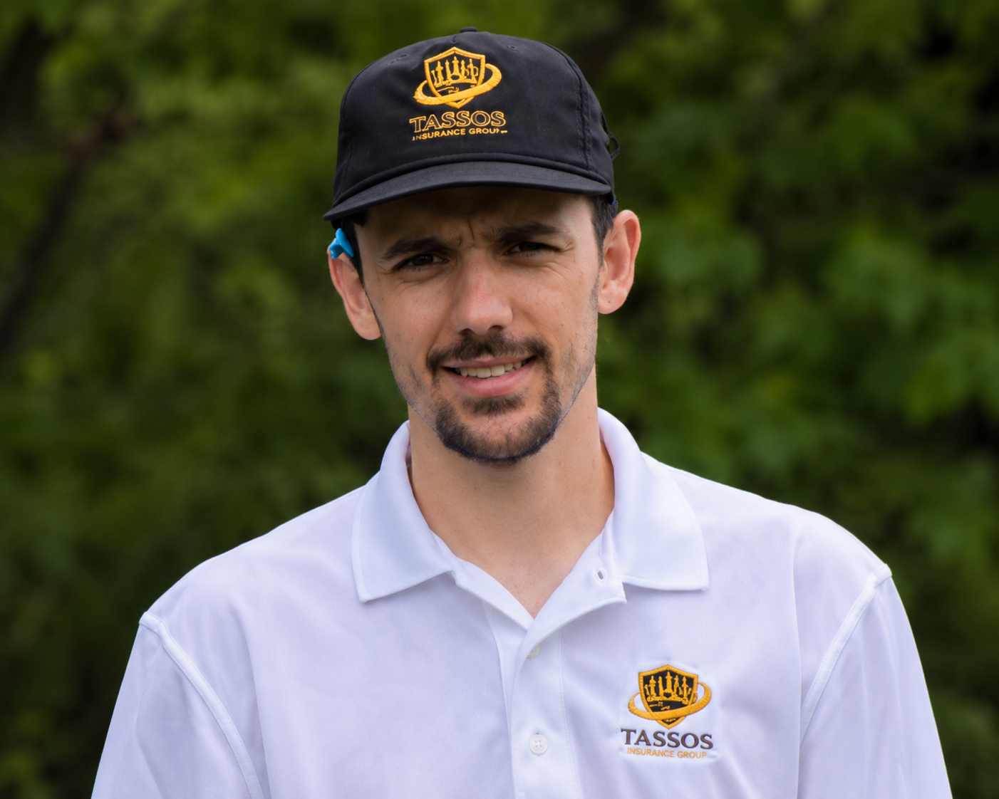 A man wearing a white polo shirt and a black cap standing outdoors with greenery in the background. The shirt and cap both display a logo for TASSOS Insurance Group.