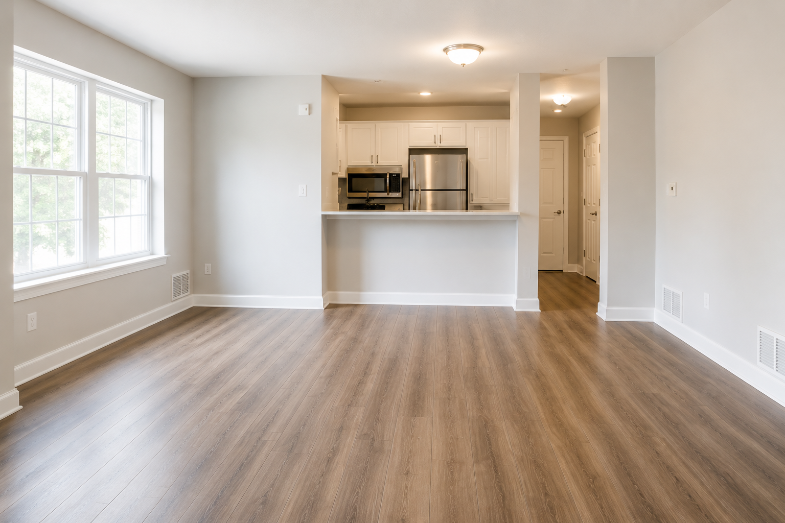 Empty living room with hardwood floors, large window, and an open kitchen with white cabinets and stainless steel appliances.