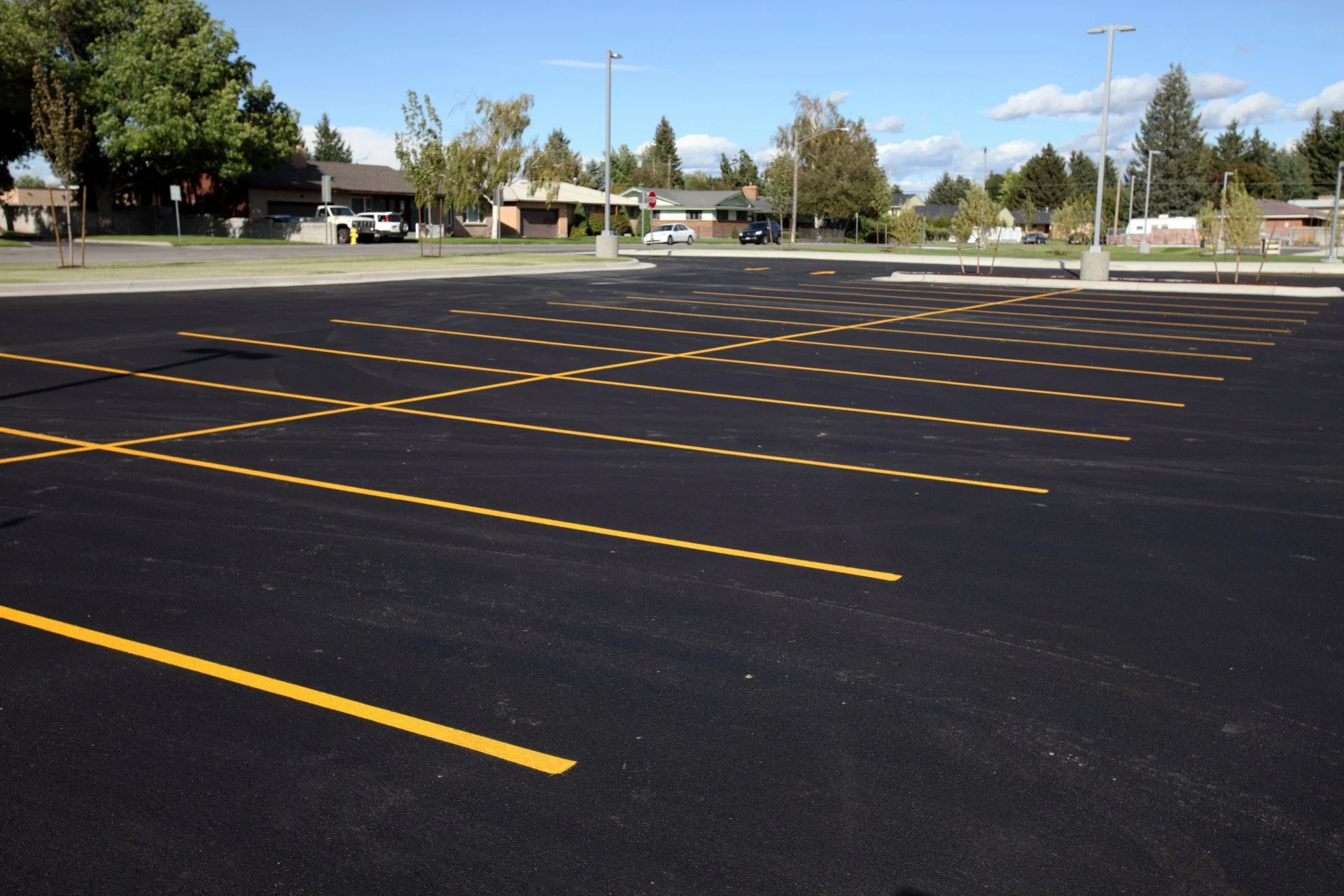 Empty parking lot with newly painted yellow lines, residential neighborhood in the background, trees, and a clear blue sky.