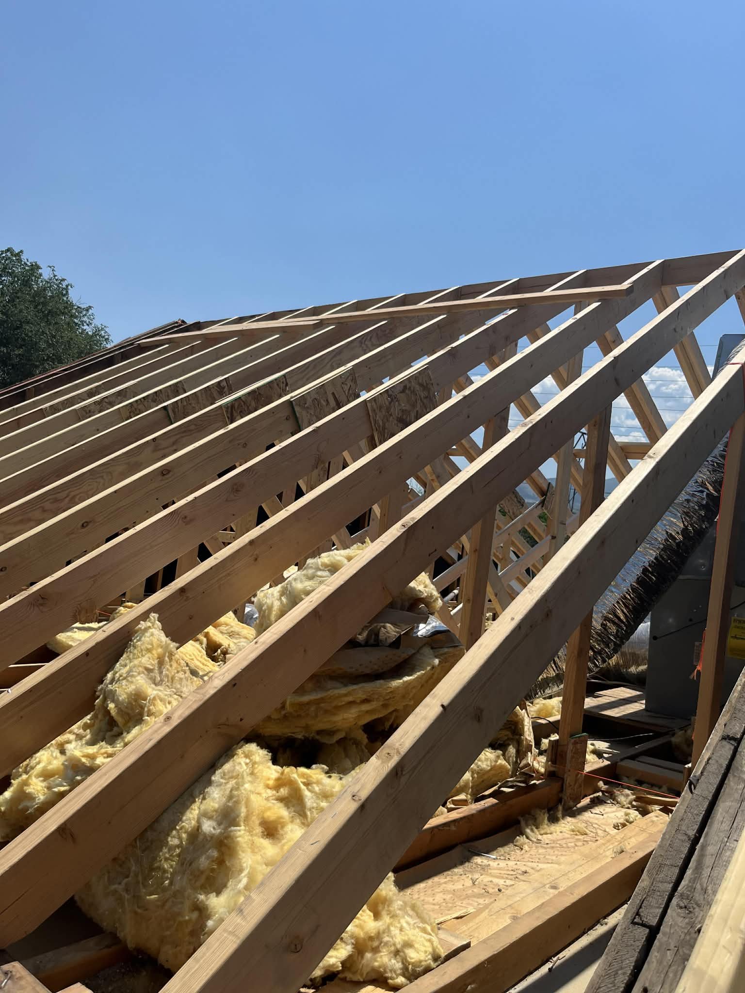 Wooden roof framing under construction with insulation material visible on the ground, clear blue sky in the background.
