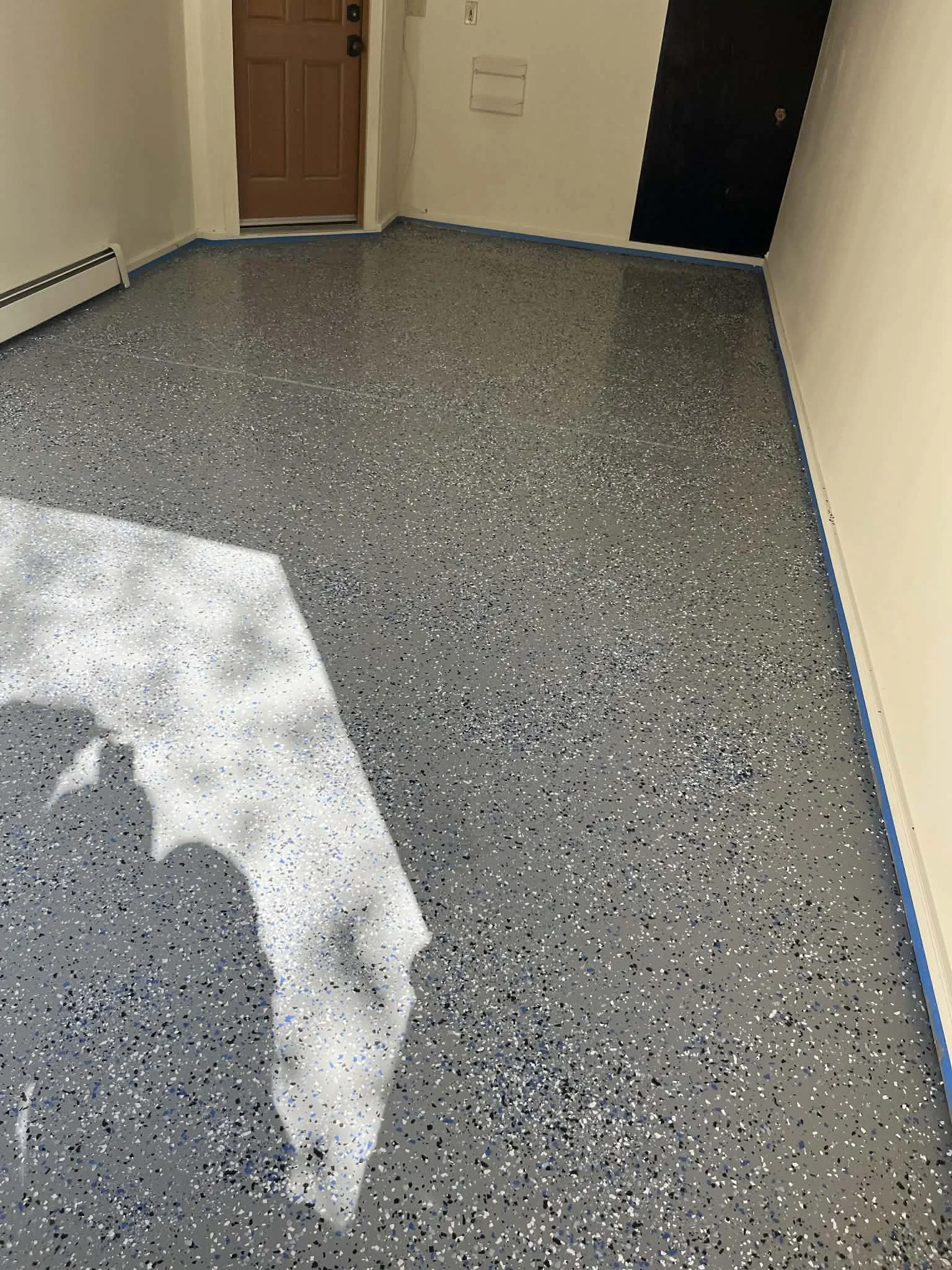 Photo of a room with a speckled epoxy floor, a closed front door, and white walls. There is a heater along the bottom left and blue painter's tape along the baseboards.
