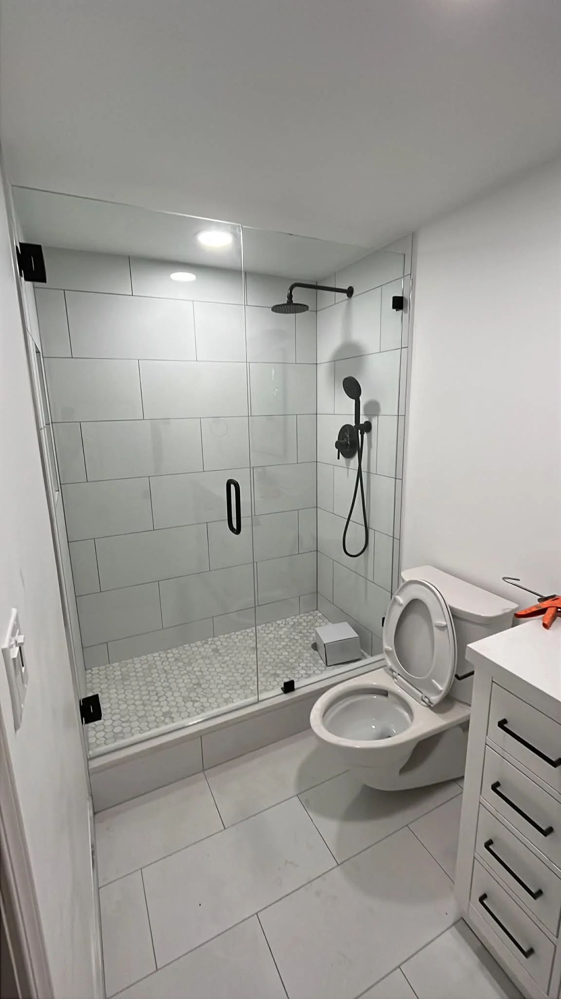 Modern bathroom with a glass-enclosed shower featuring black fixtures, a white toilet, and a white vanity with drawers. The shower has large white tiles and a small hexagon-tile floor. The ceiling has recessed lighting and white walls.