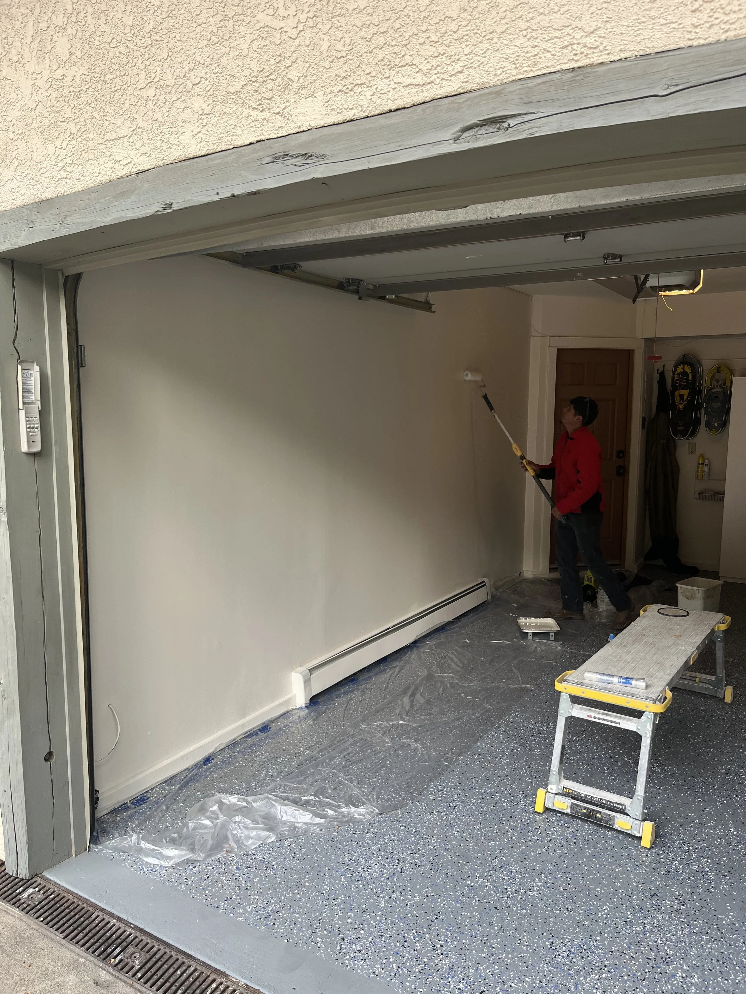 Person painting the interior garage wall with a roller in a residential home, with painting supplies and tools around.