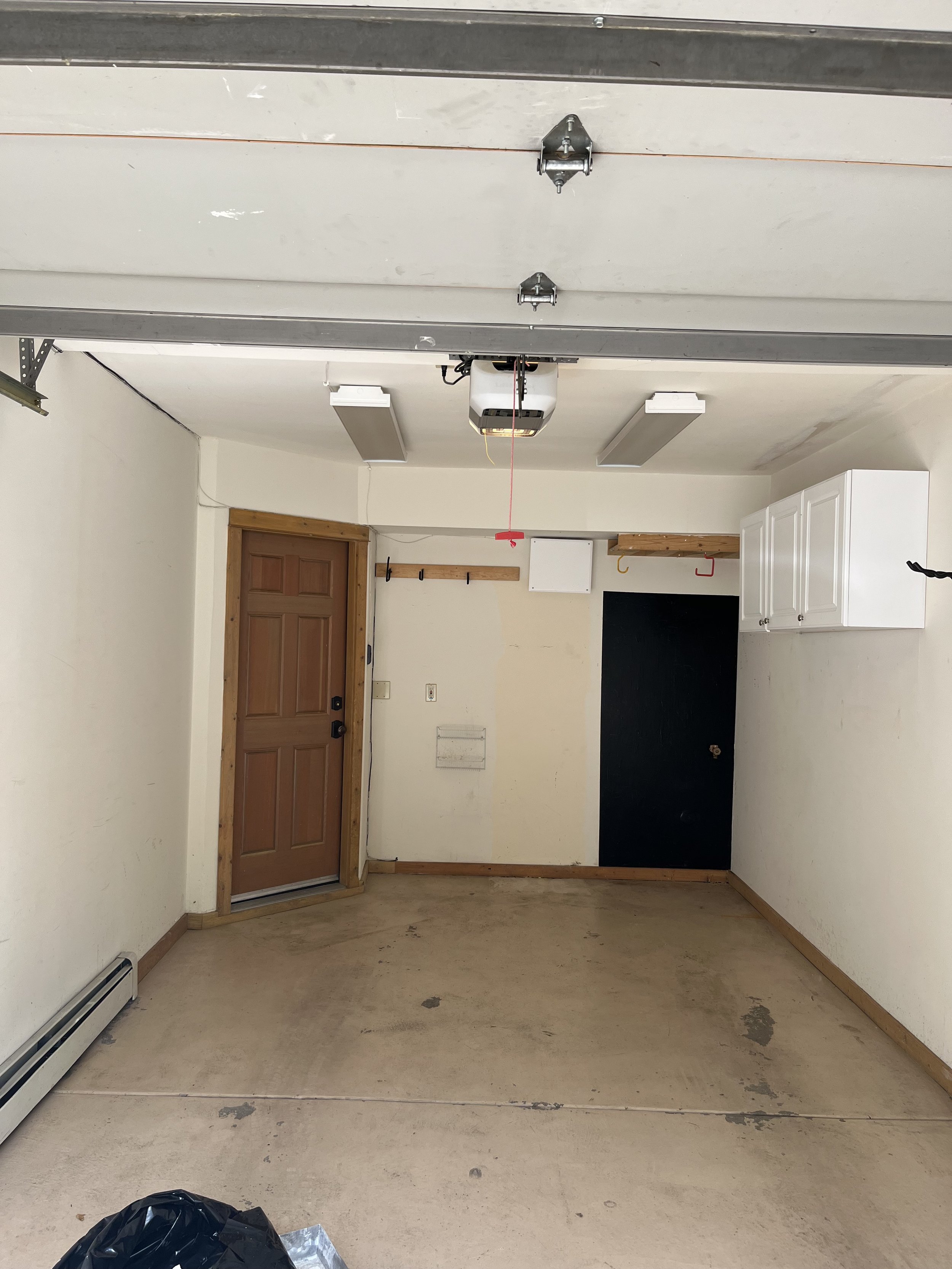 An empty garage with a black floor and white walls, a wooden door, utility hooks, and some trash on the floor.