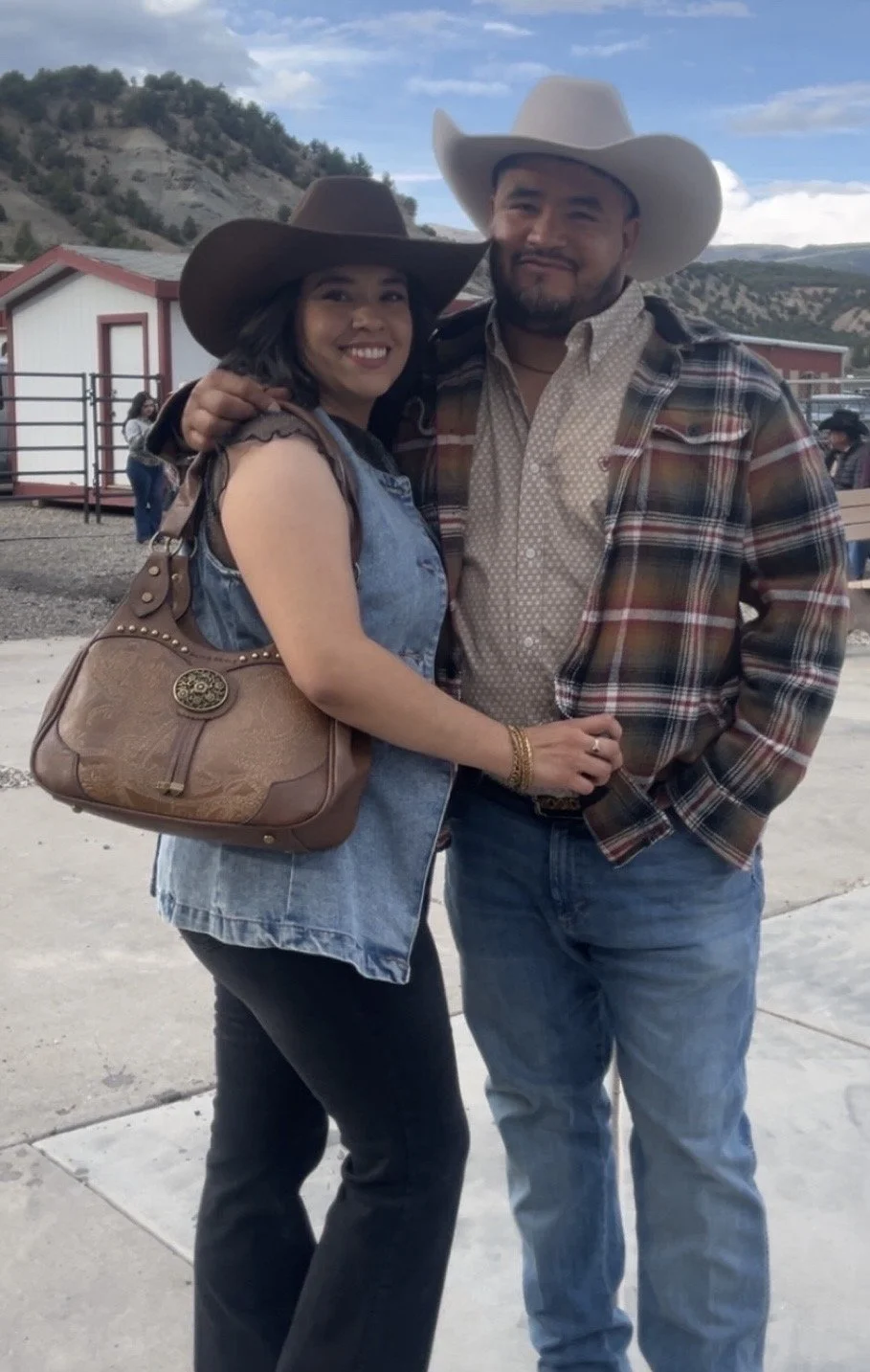 A smiling woman and man wearing cowboy hats, embracing outdoors on a cloudy day with mountains and a red and white building in the background.