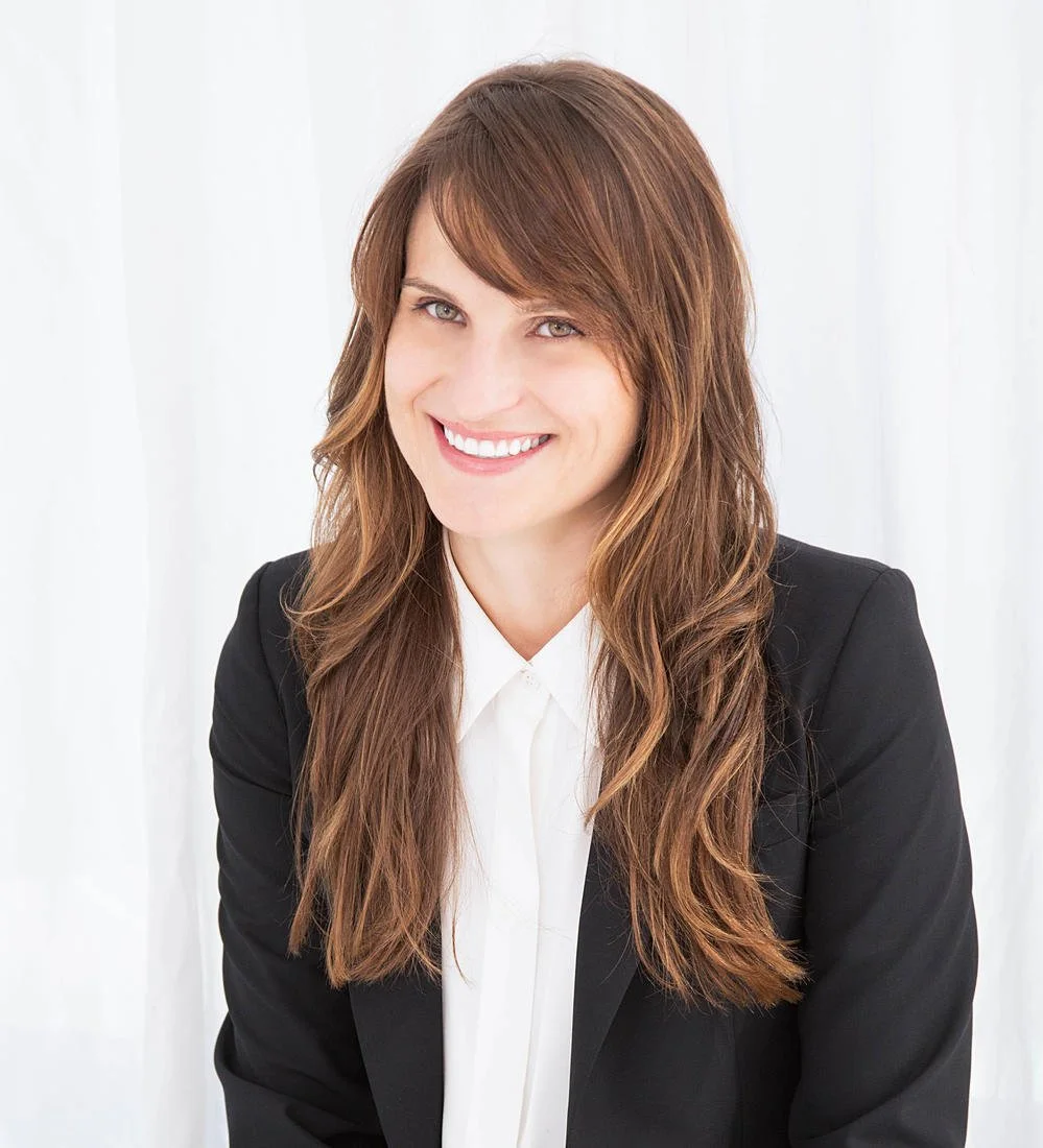 Headshot of Dr. Vogel, a white woman with long brown wavy hair, smiling at the camera. She is wearing a professional white blouse under a black blazer and seated in front of a white backdrop.