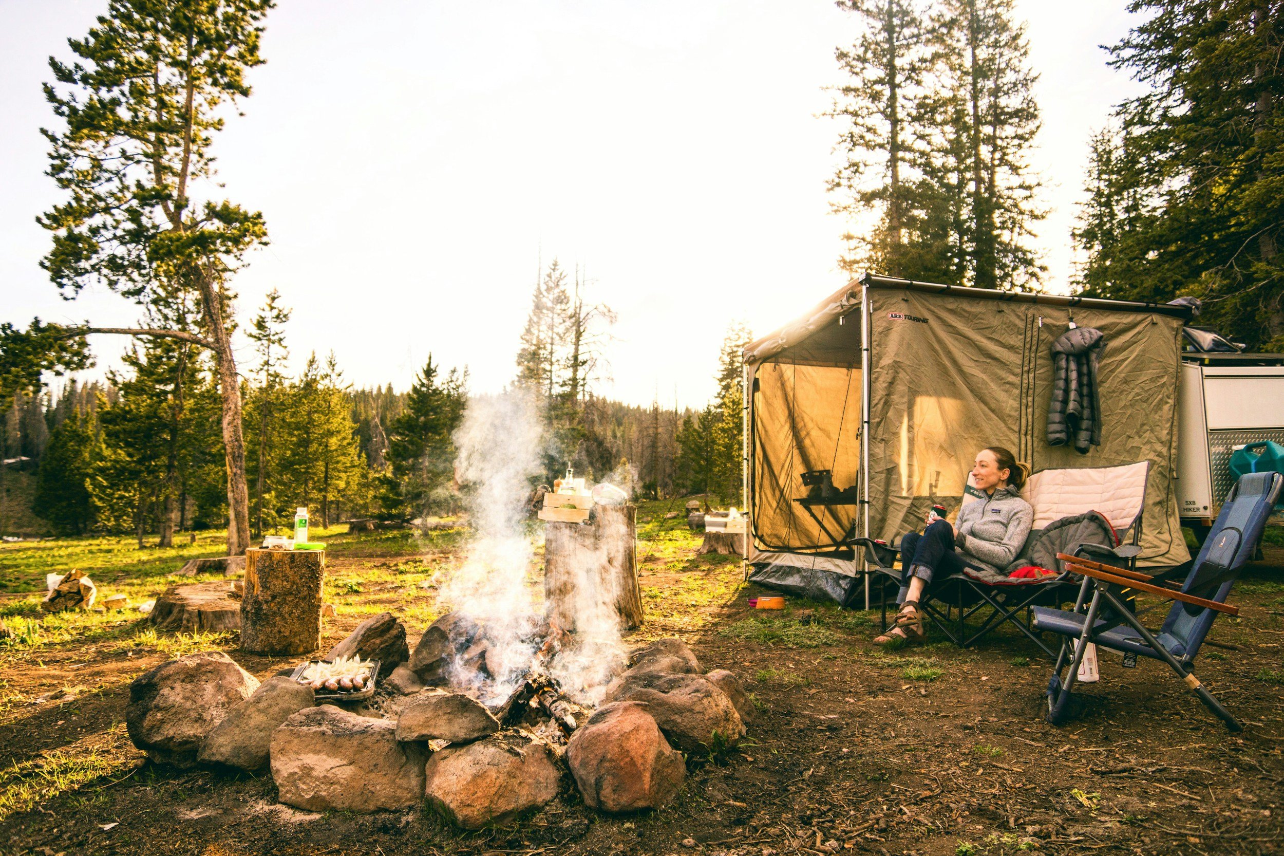Person sitting in camping chair near a campfire in a forested area, with a canopy tent and trees in the background