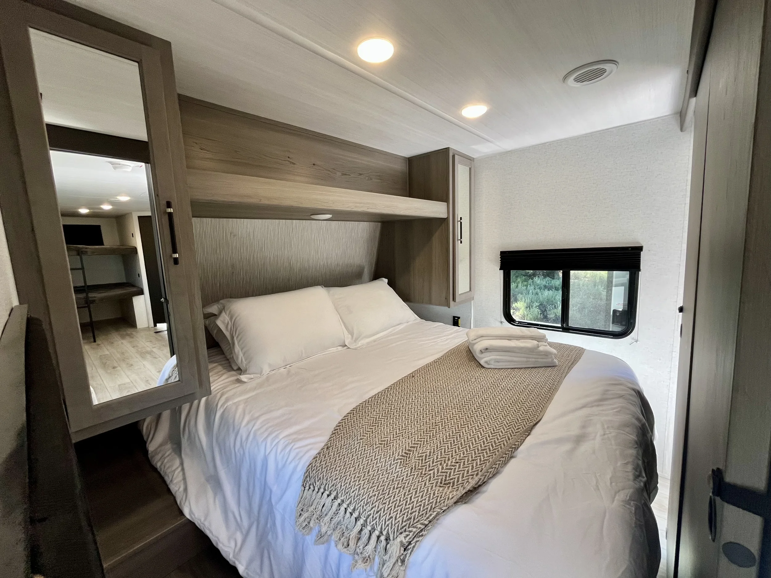 A bedroom with a bed covered in white bedding, a beige patterned blanket at the foot of the bed, a small window with black blinds, wooden furniture including a headboard with built-in shelves, mirrors, and cabinets, and ceiling lights.