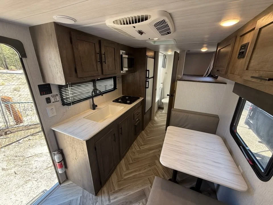 Interior of an RV kitchen with a sink, stove, microwave, refrigerator, small dining table with benches, and a view of a bathroom.